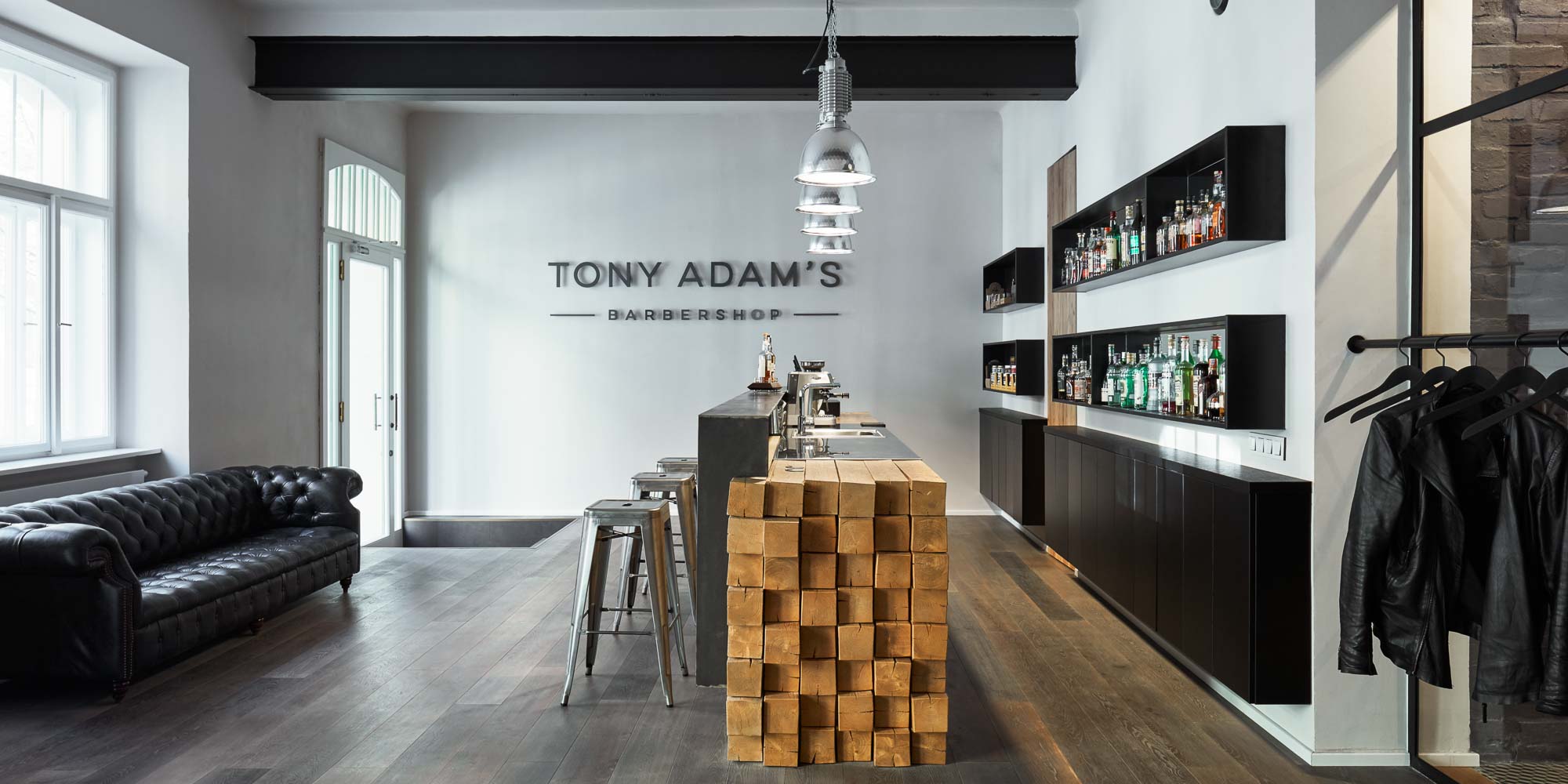 Celkový pohled na vstupní prostor barber shopu Tony Adam's od OOOOX – bar z masivních dubových trámů s ocelovým pultem, černá kožená Chesterfield sedačka, police s lihovinami a industriální závěsná světla