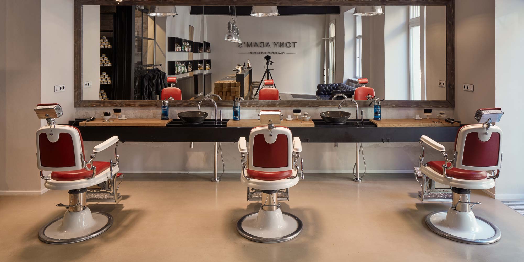 Tři retro holičská křesla v červené kůži v barber shopu Tony Adam's – ocelová traverza s integrovanými umyvadly a dubovou policí na kosmetiku, velkoformátové zrcadlo s odrazem vstupní části a logem