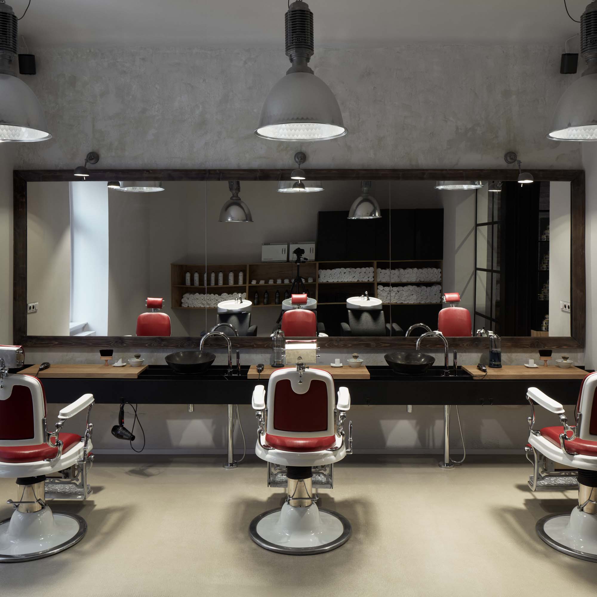 Holičská stanoviště v barber shopu Tony Adam's – tři retro křesla v červené kůži před velkým zrcadlem v dřevěném rámu, ocelová traverza s integrovanými umyvadly, škrábaná omítka na stropě a industriální závěsná světla