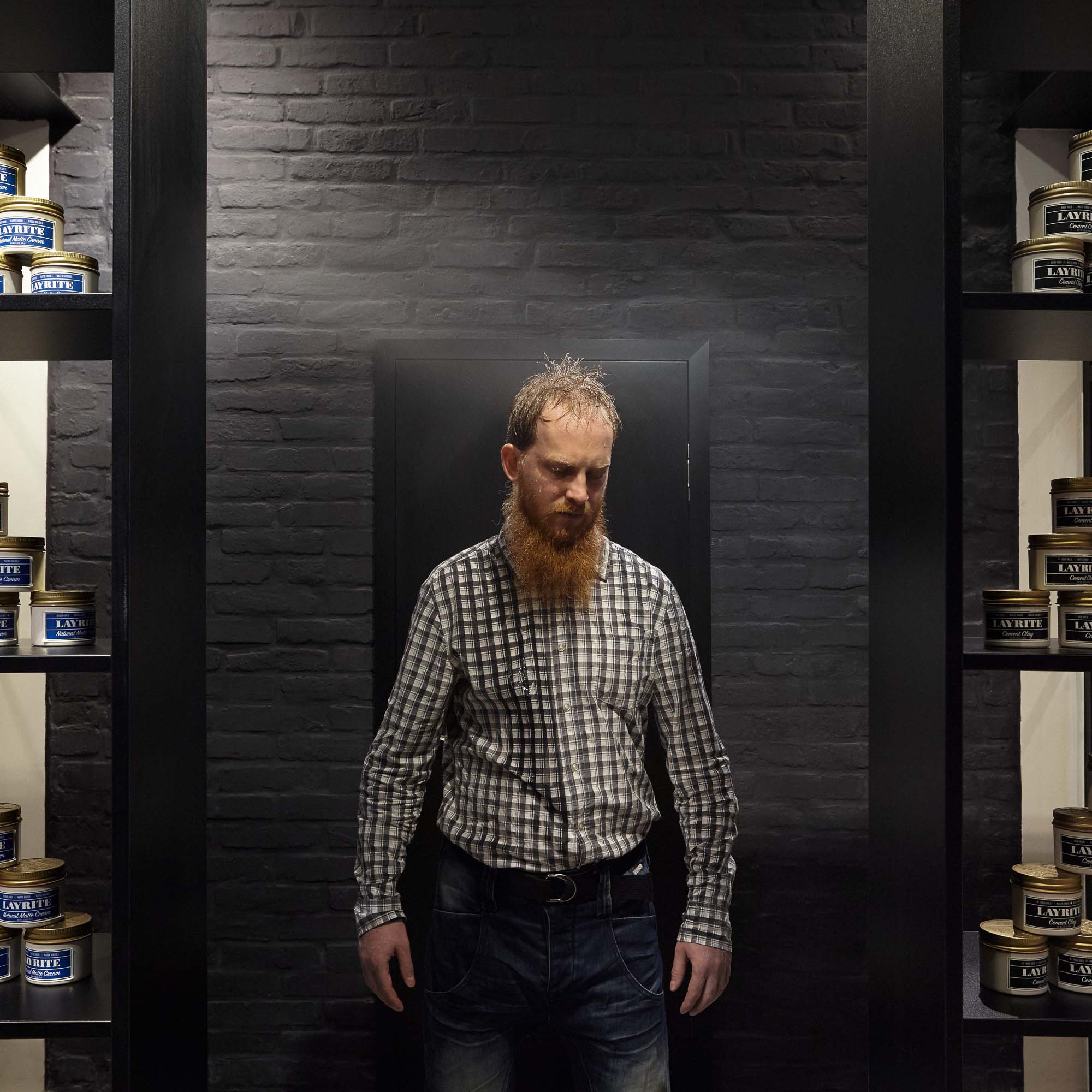 Muž s rudým vousem v produktovém koridoru barber shopu Tony Adam's – černé ocelové regály s pomádami Layrite po obou stranách a odkrytou natřenou cihlovou stěnou za zády