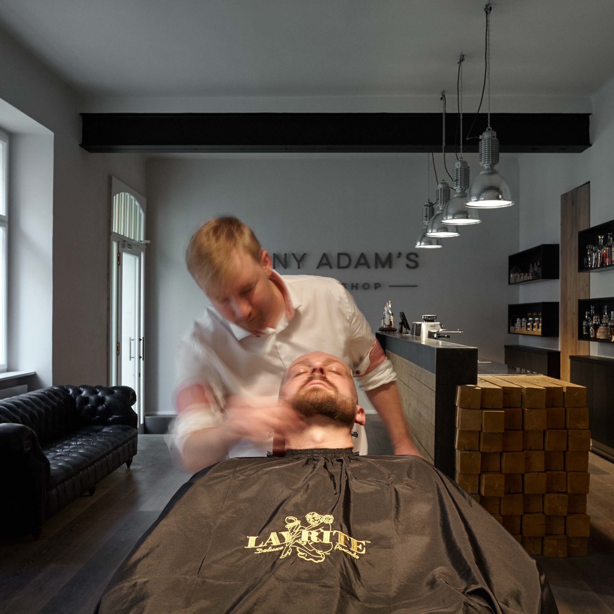 Barber při ošetření zákazníka v holičském křesle barber shopu Tony Adam's – zákazník s pláštěm Layrite v červeném retro křesle, v pozadí vstupní prostor s logem a barem