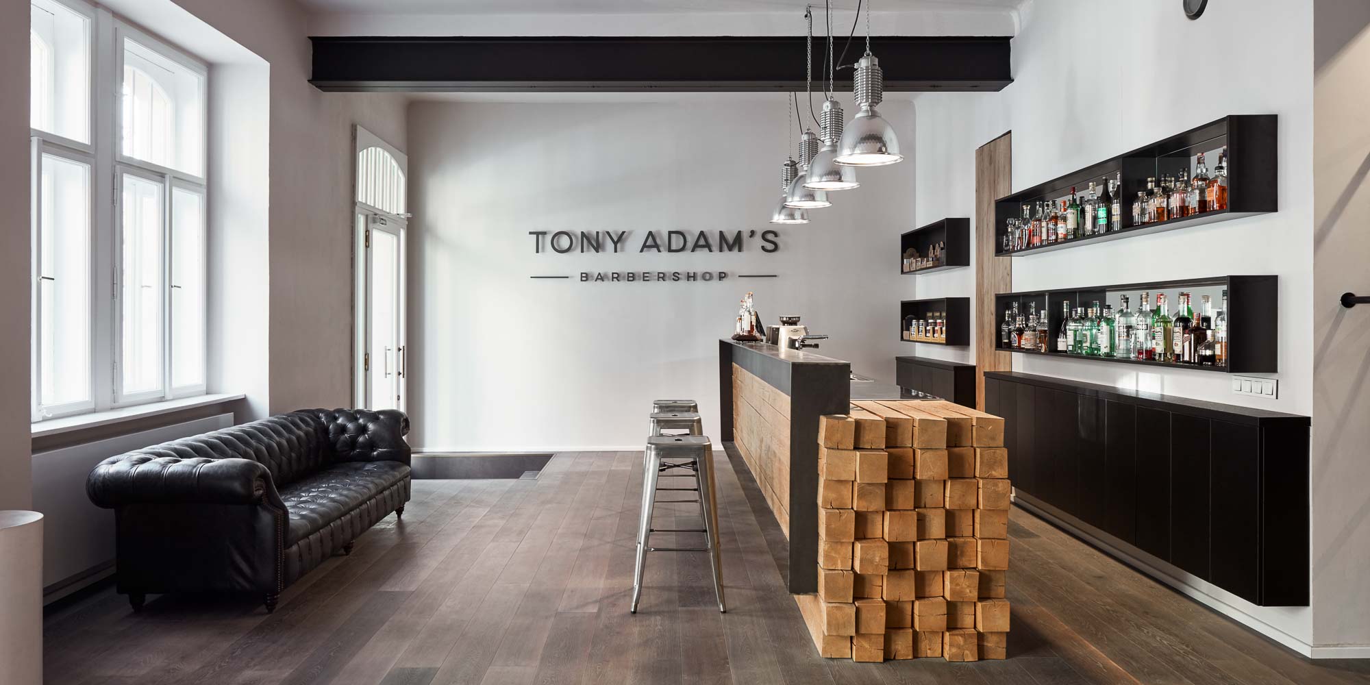 Vstupní prostor barber shopu Tony Adam's s logem na stěně – černá kožená Chesterfield sedačka, bar z dubových trámů se stoličkami, ocelové police s lihovinami a industriální závěsná světla