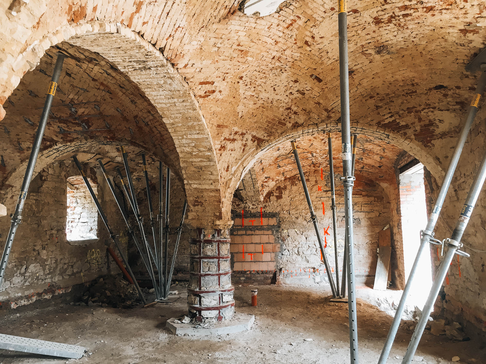 Rekonstrukce historického domu — stabilizace cihelných valených kleneb ocelovými vzpěrami a lešením, pohled na trojici klenebních polí s poškozenou omítkou a sprejmovanými pracovními značkami