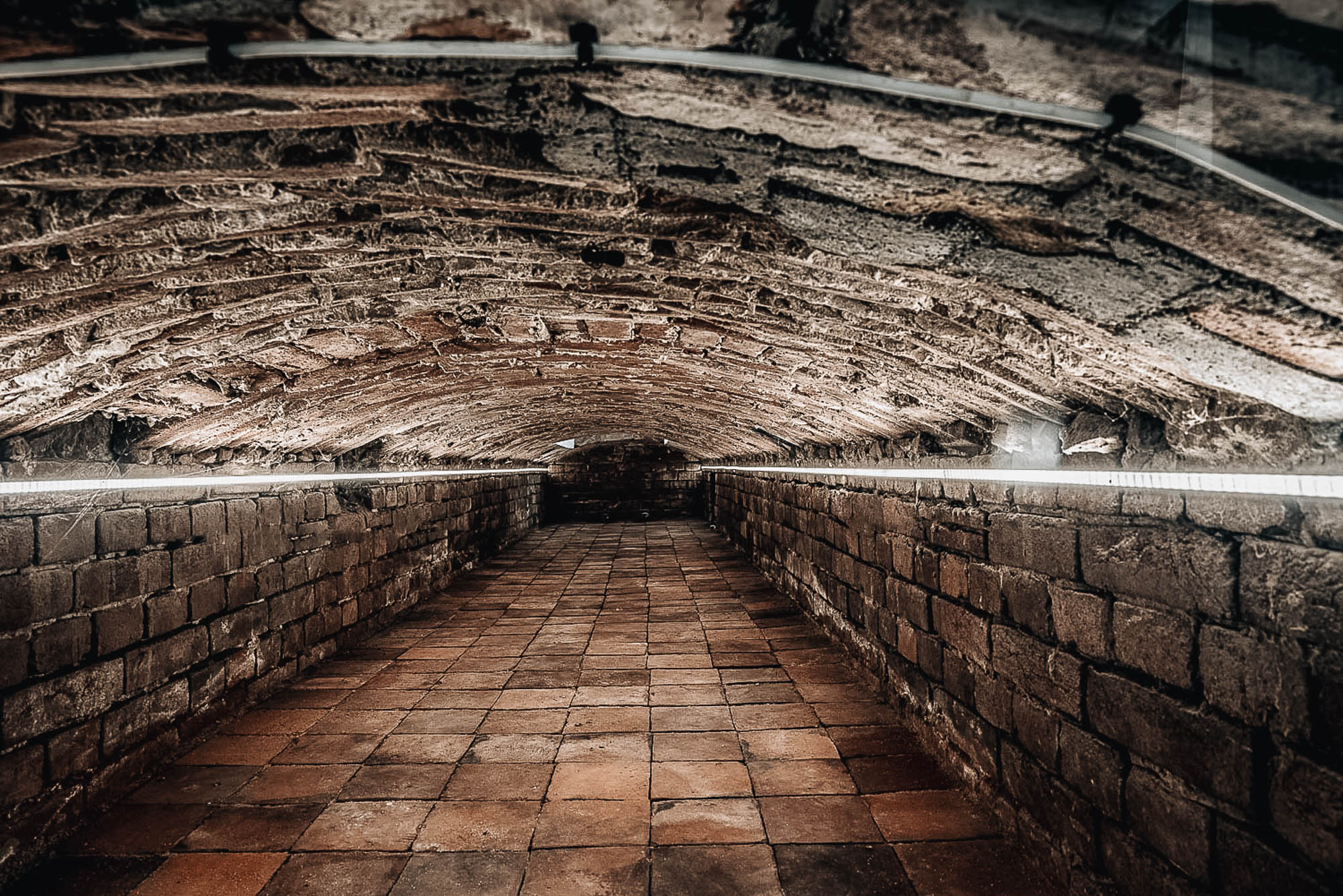 Podzemní chodba historického domu s valenou klenbou — dlouhý klenutý tunel s terrakotovou dlažbou, hrubé kamenné zdivo osvětlené lineárními LED pásky, dramatická perspektiva do hloubky prostoru