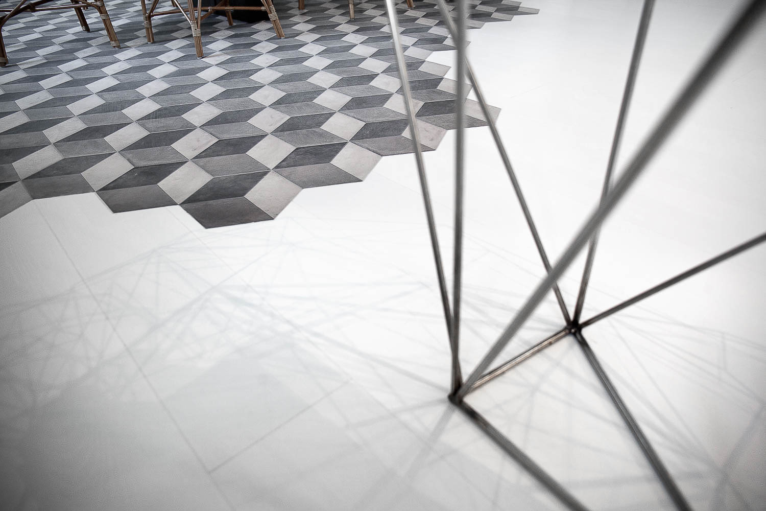 Floor combination detail: geometric hexagonal tiles in grey and white tones transitioning into smooth white concrete screed — metal table leg in foreground, wicker garden chairs in background