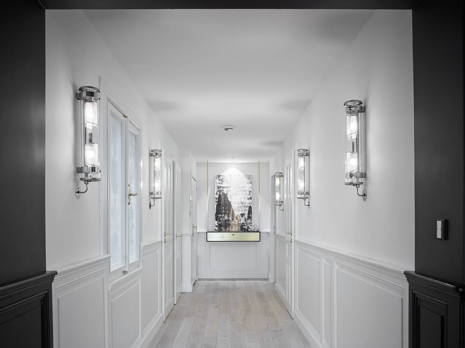 White statement corridor with dark-painted side walls and white panel moulding along both sides — chrome wall sconces flanking the passage, light wooden floor, abstract artwork at far end
