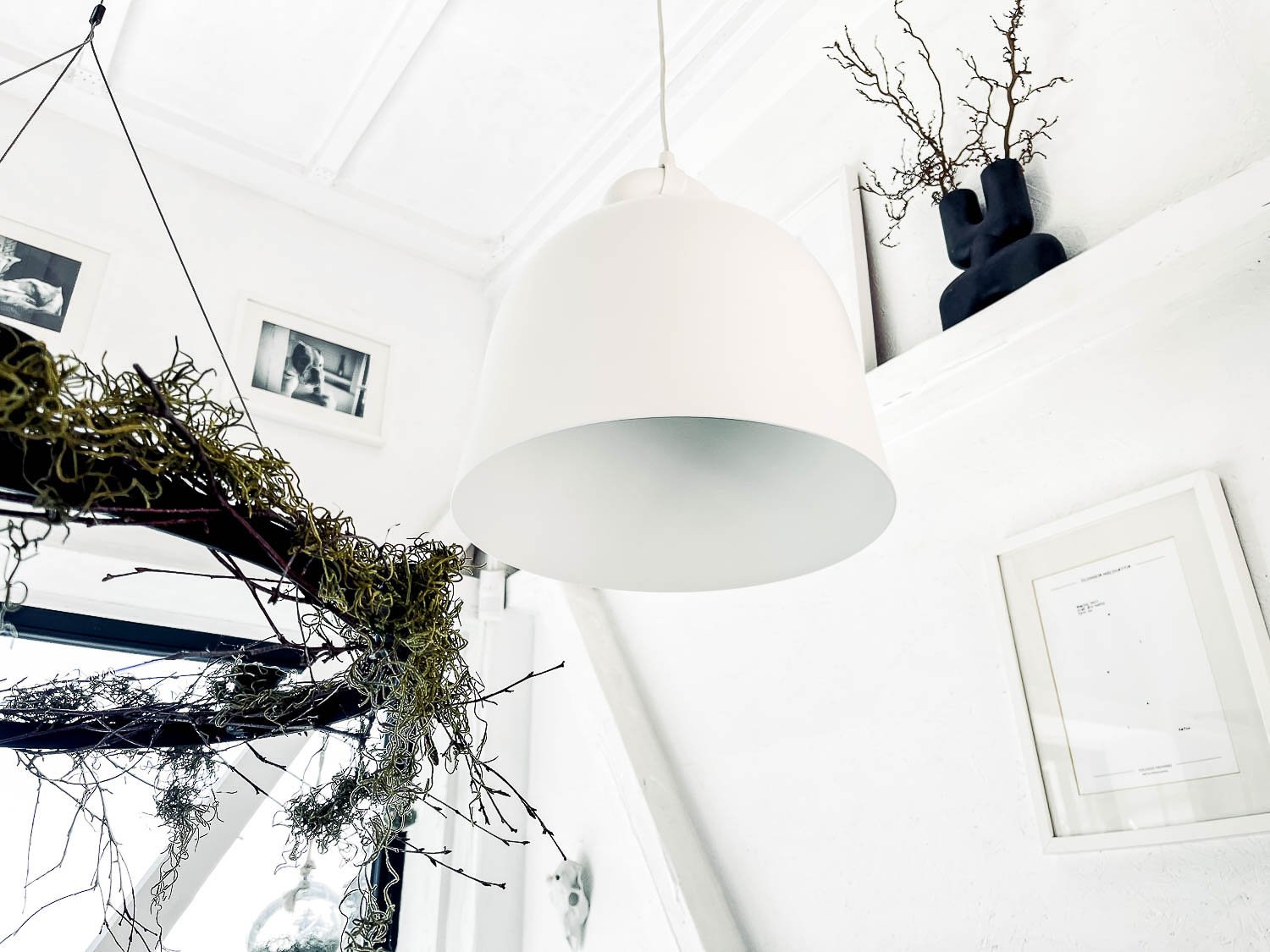 View from below of white bell pendant lamp in white attic interior — decorative dry branches and black vase on white shelf, framed artwork; inspiration for white Scandi interior