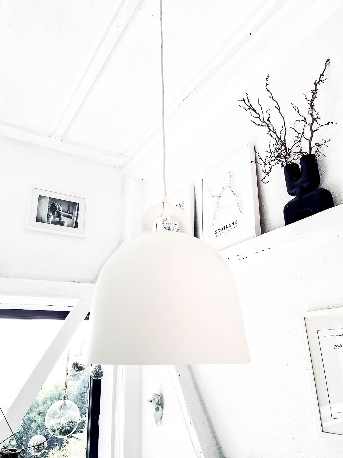 View from below of white bell pendant lamp in white attic interior — decorative dry branches and black vase on white shelf, framed artwork; inspiration for white Scandi interior
