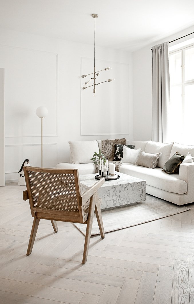 Scandinavian living room in all-white — rattan cane armchair in foreground, white corner sofa, marble coffee table, waxed pine herringbone floor and multi-arm chandelier