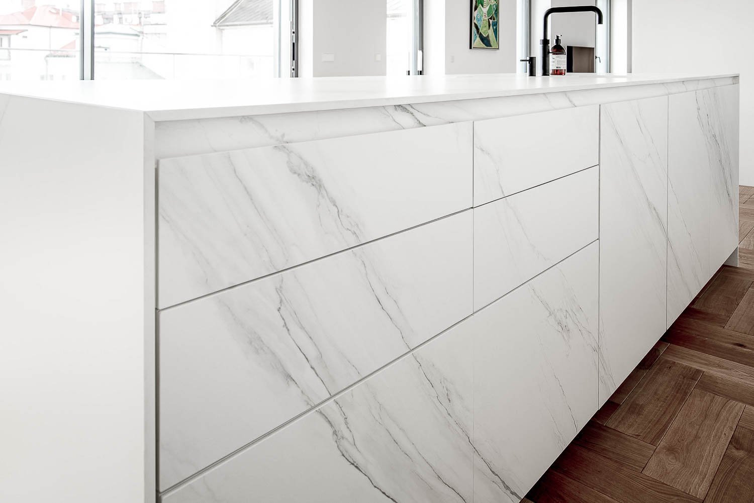 Detail of kitchen island with front panel in white veined sintered stone — thin-edge worktop, herringbone parquet floor, large windows in background