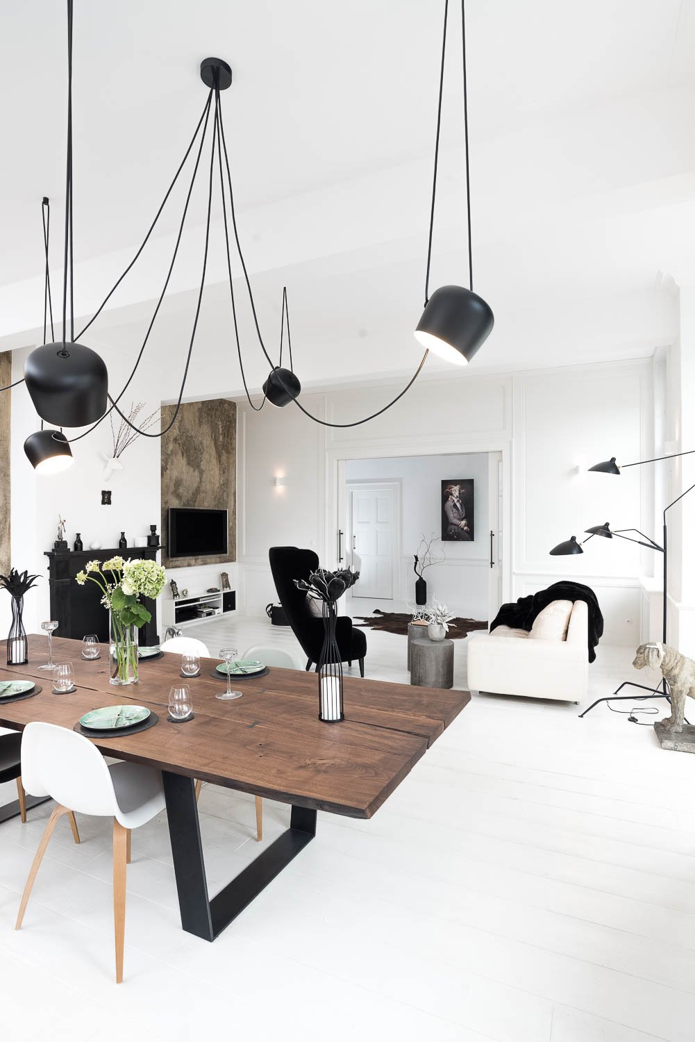 Generous white apartment with dark solid-wood dining table with black patina, multi-arm pendant lamps (Flos Aim), white sofa and gallery artwork — contrast of dark timber and white space