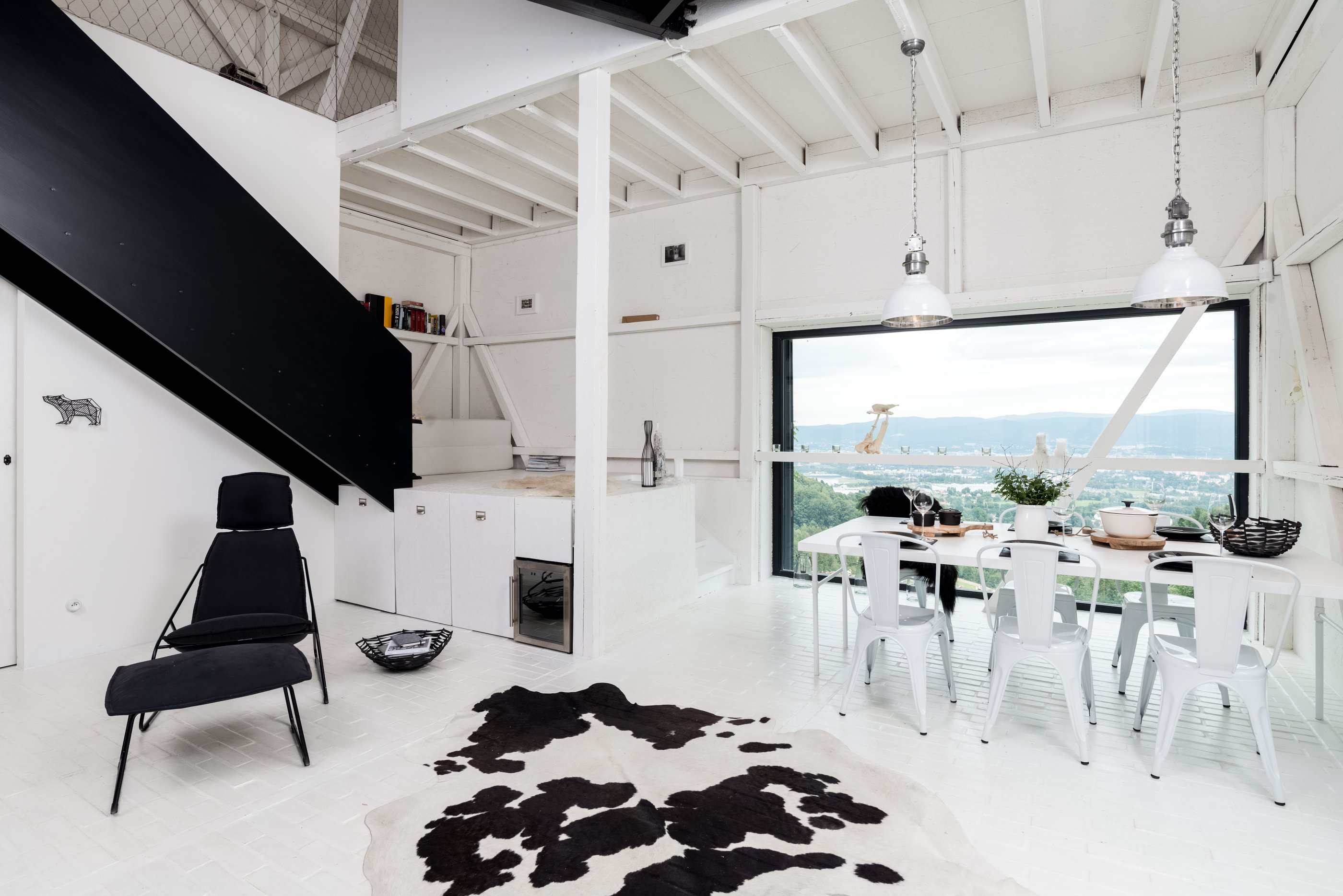 White loft living space with black steel staircase — black-and-white cowhide rug, white dining table with metal Tolix chairs, industrial pendant lamps and panoramic window with landscape view