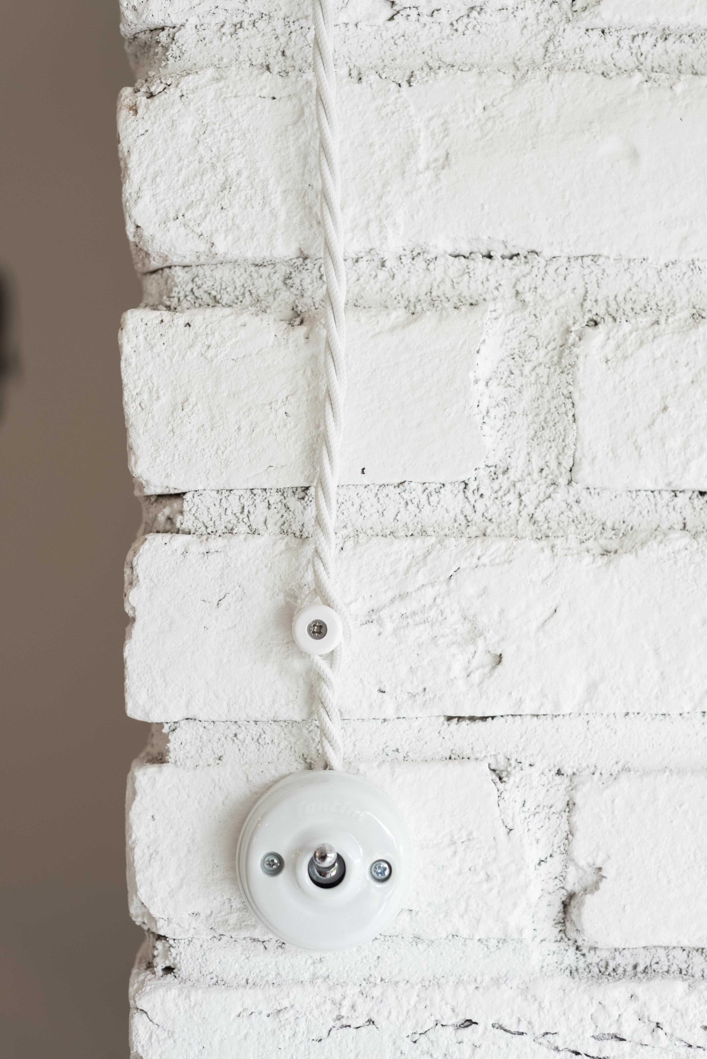Detail of white porcelain rotary light switch with twisted textile cord on corner of white painted brick wall — surface contrast in a white industrial interior