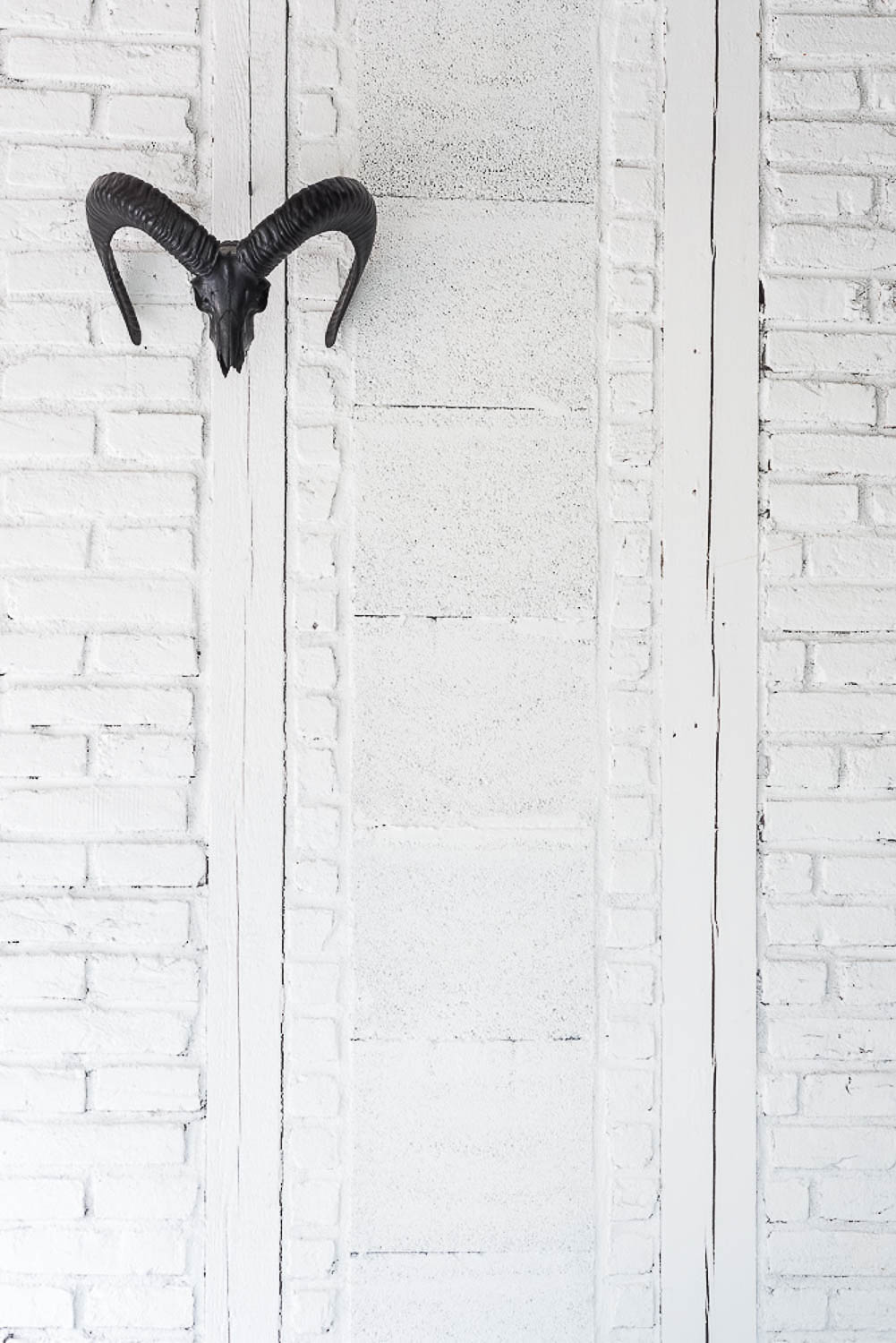 Black metal ibex skull mounted on white painted brick wall — decorative accent emphasising the texture of white-painted brick masonry in an industrial white interior