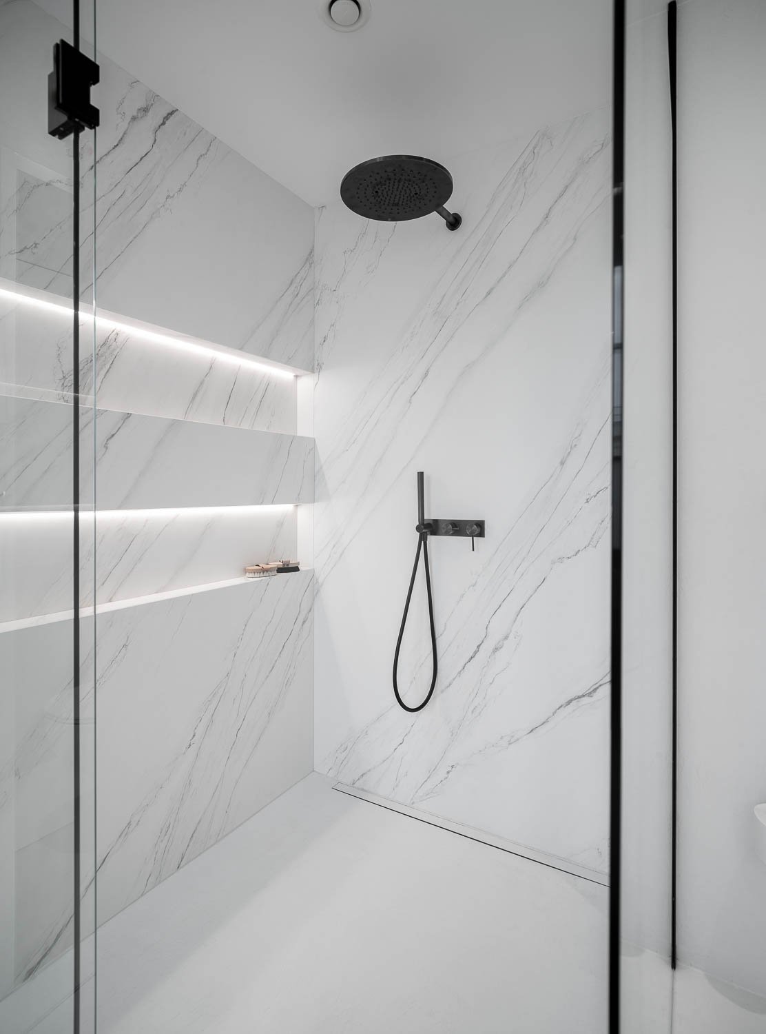 White marble shower enclosure with black overhead rain shower and hand shower, black thermostatic module, LED backlit niche shelf with toiletries and glass shower screen