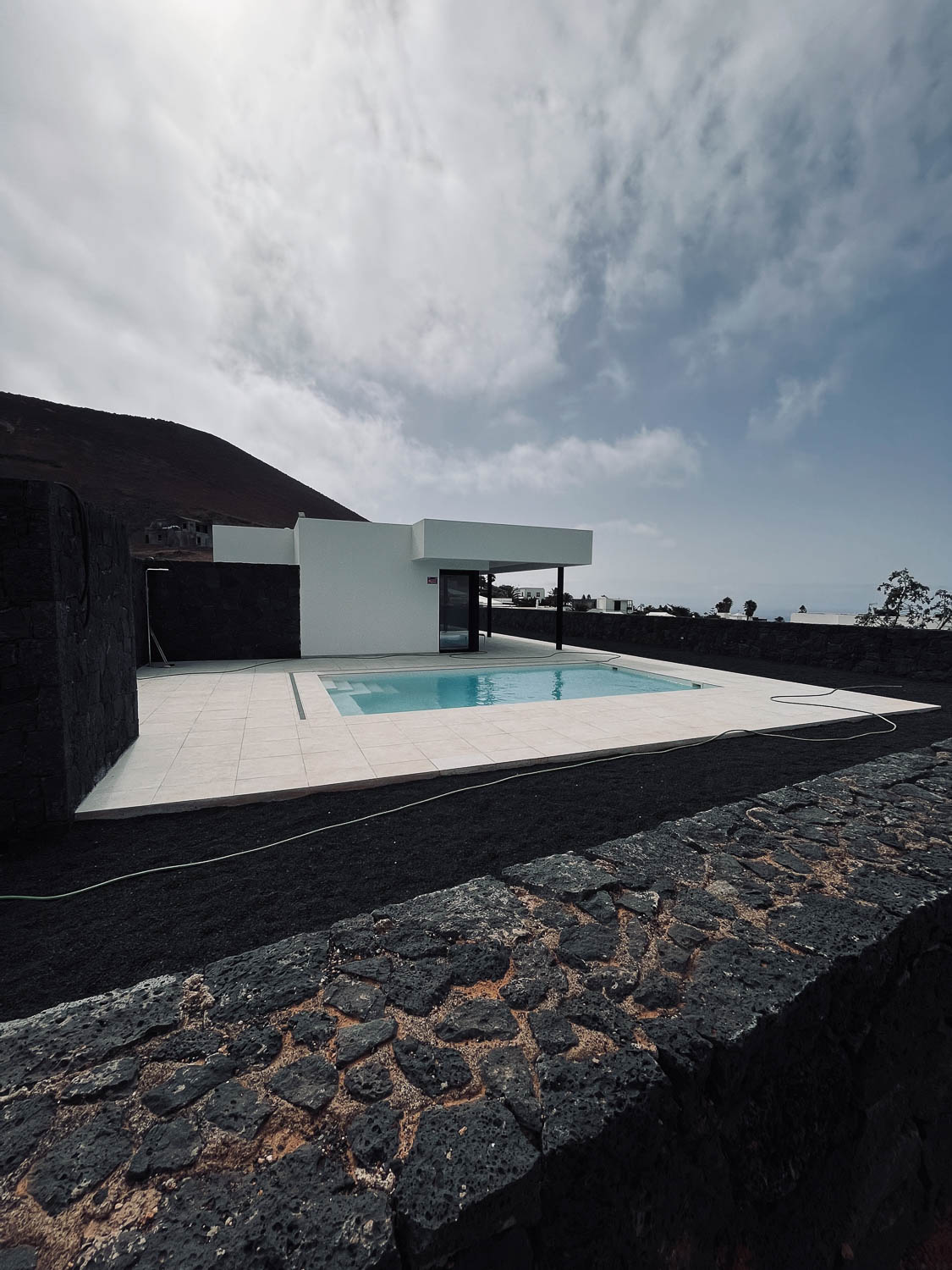 Minimalistická bílá vila s obdélníkovým bazénem a lávovou kamennou zdí v popředí, výhled na oceán, Lanzarote