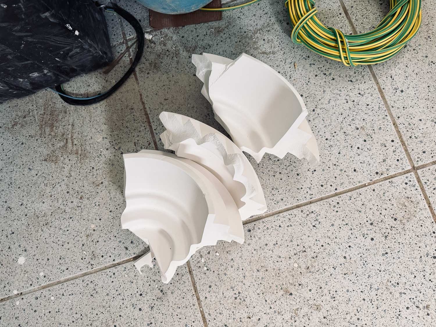 Rozbité bílé keramické svítidlo rozpadlé na kusy na dlážděné podlaze vedle elektrického kabelu