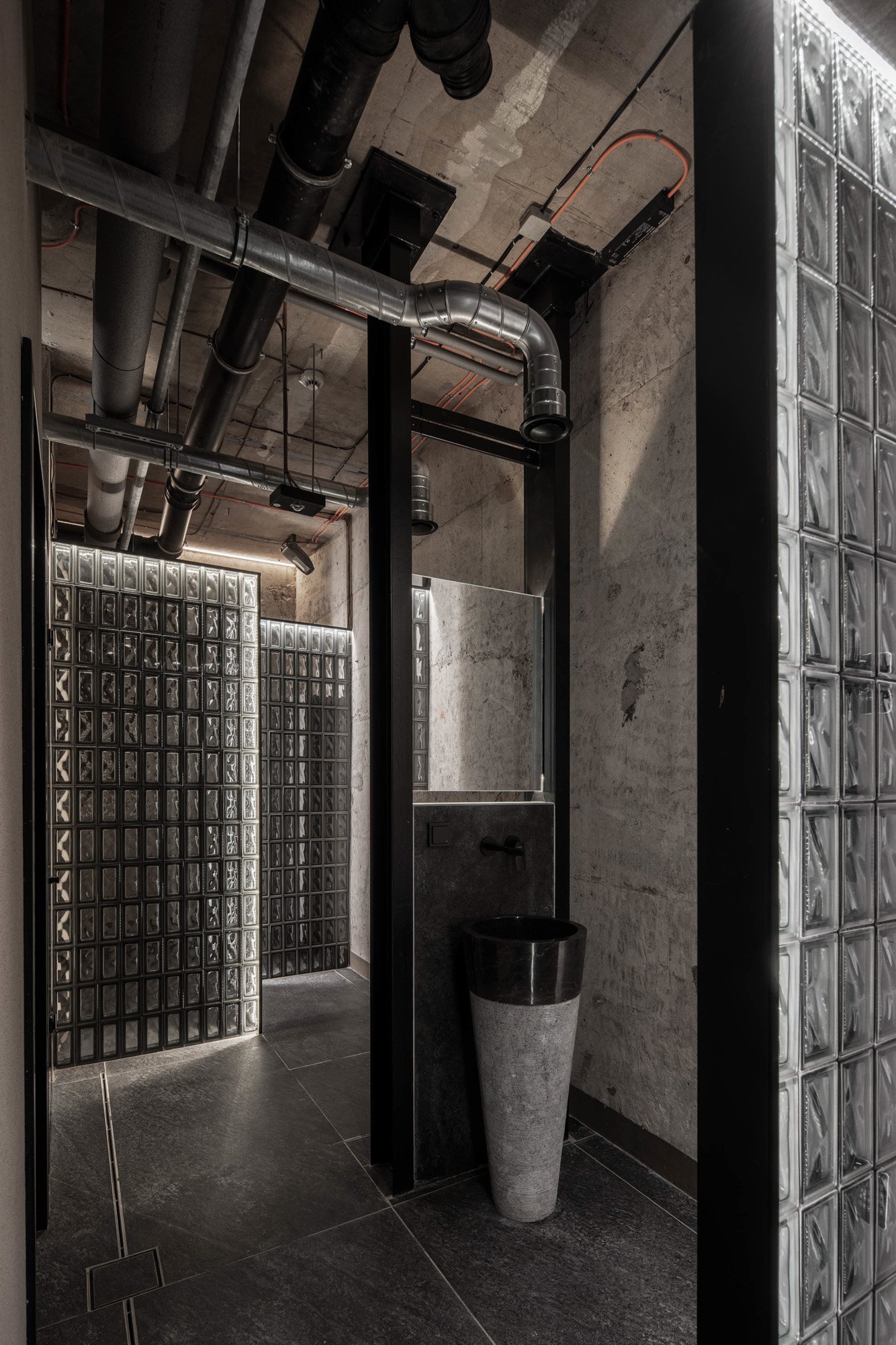 Industriální prostor — příčka ze skleněných luxfer, černá crittall a bílé keramické sloupcové umyvadlo