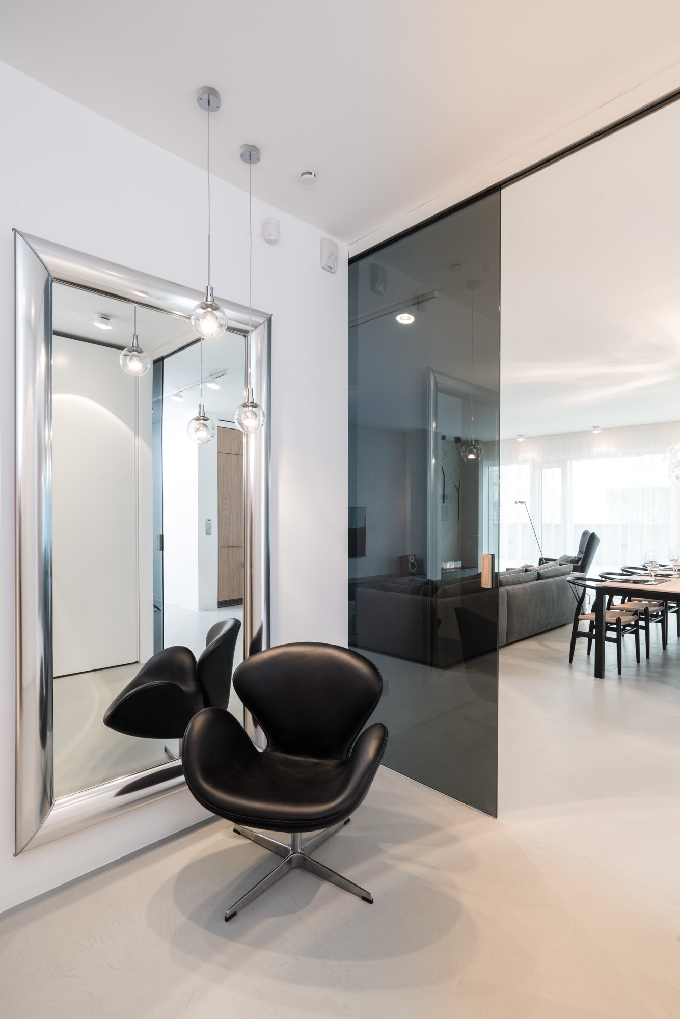 Bílý minimalistický interiér s černou crittall příčkou — tmavé designové křeslo v popředí a závěsná světla