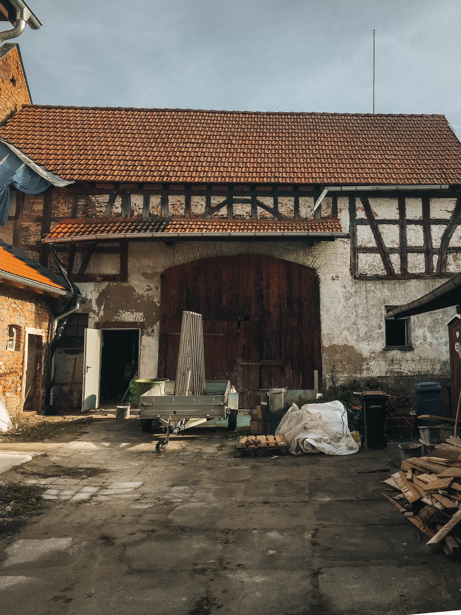 Fachwerková stodola před rekonstrukcí — velká oblouková vrata, omšelá fasáda s pohledovým hrázděním a stavební materiál na dvoře