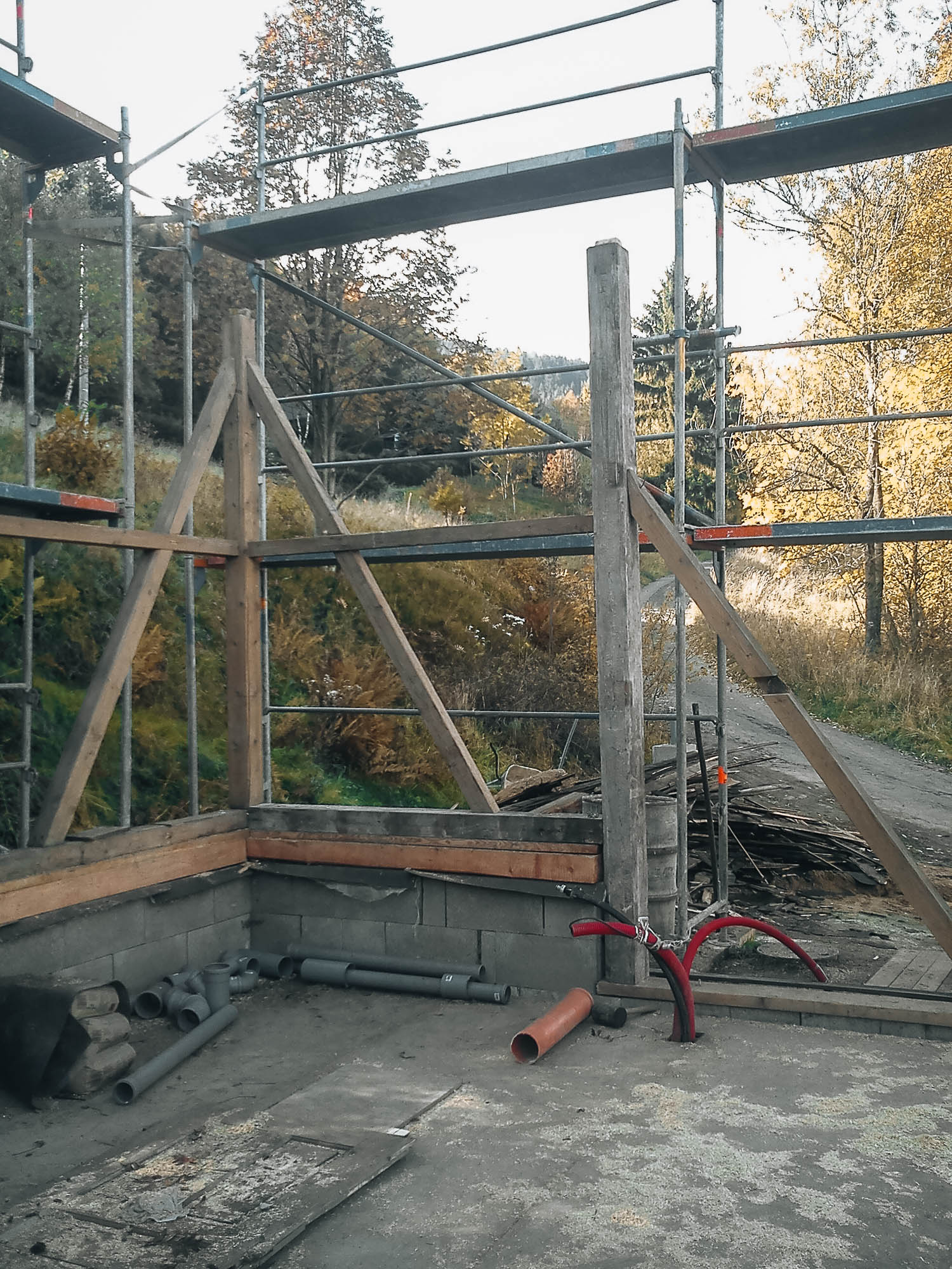 Stavba rekonstrukce stodoly — lešení se zachovaným původním dřevěným sloupem in situ, nové kanalizační trubky na betonové desce a podzimní stromy