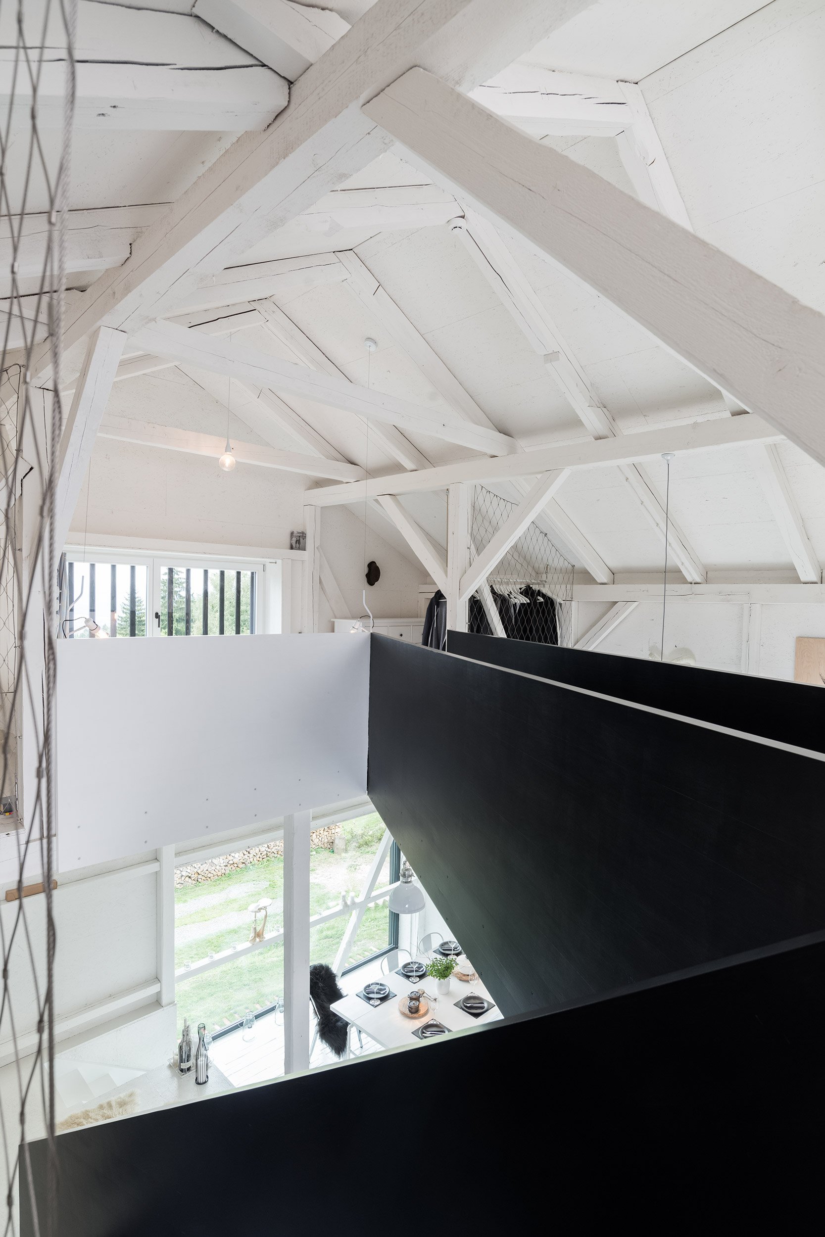 Pohled z galerie rekonstrukce stodoly BOX — bílý pohledový krov shora, hrana černého ocelového mezaninu v popředí a pohled dolů na jídelnu s panoramatickým oknem do zahrady