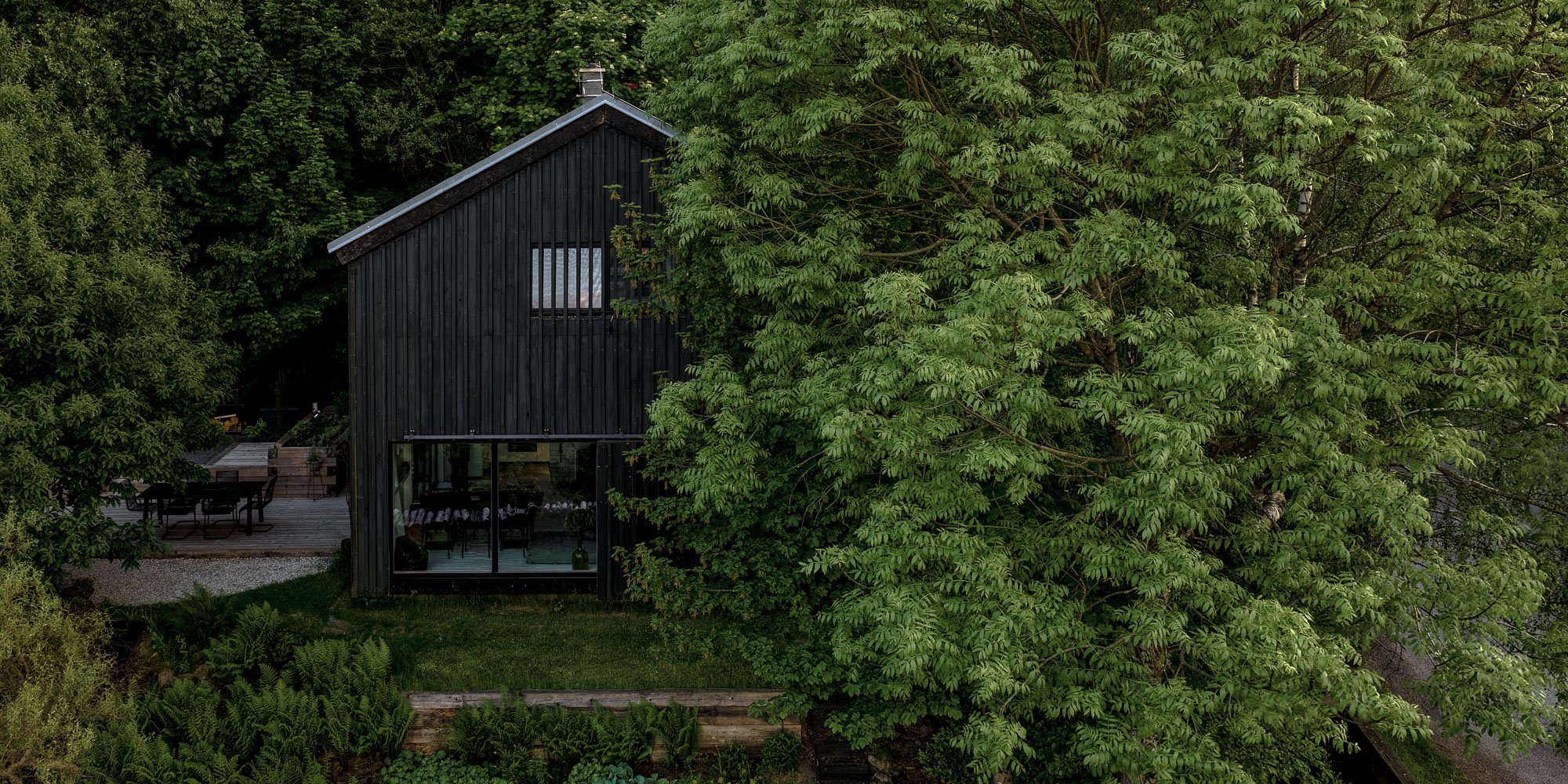 BOX barn conversion — black vertical timber cladding, large panoramic window, garden slope with stacked logs, pool on the slope | OOOOX