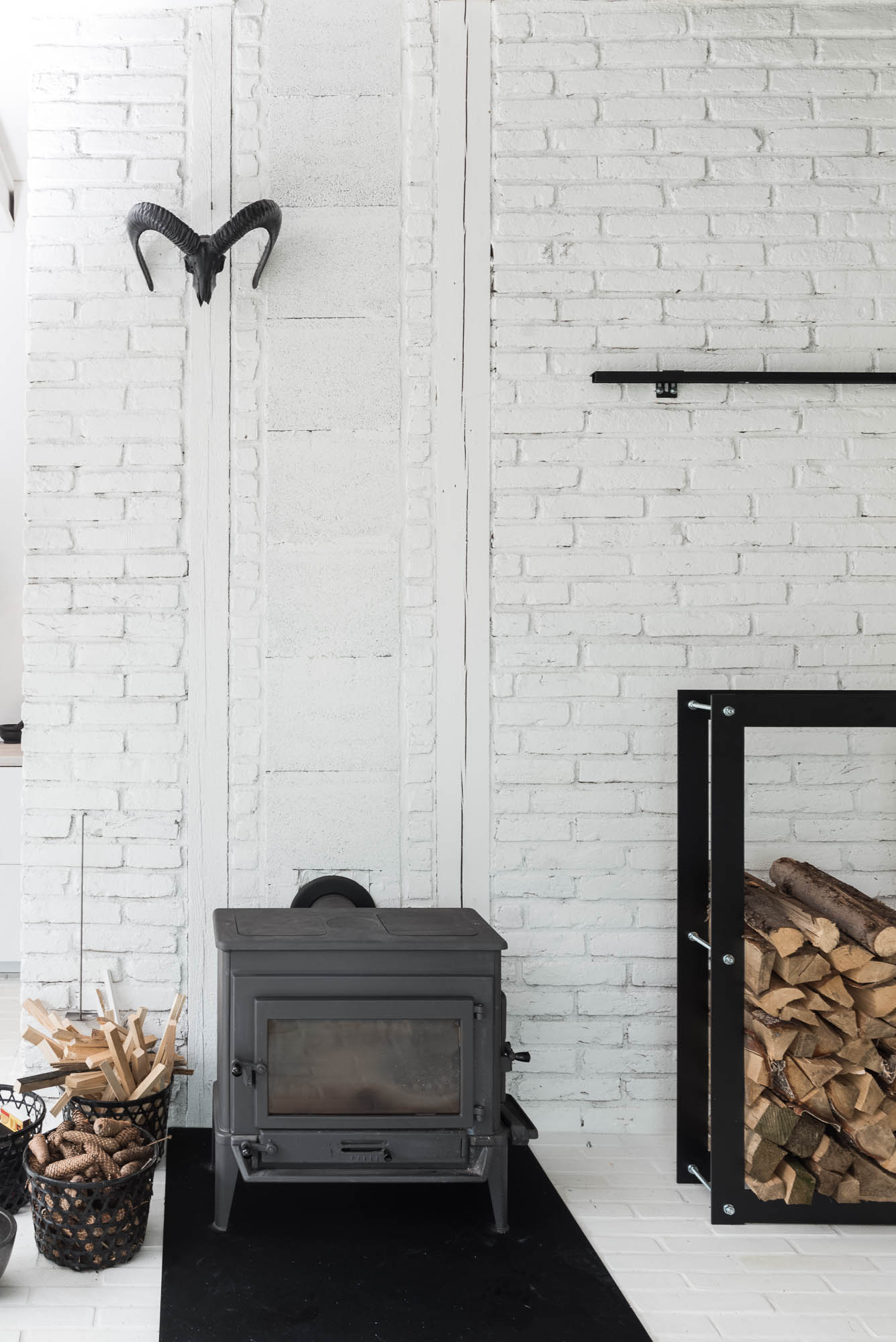 BOX barn wood stove detail — black cast iron stove, firewood in black rack, ram skull mounted on white painted brick wall | OOOOX