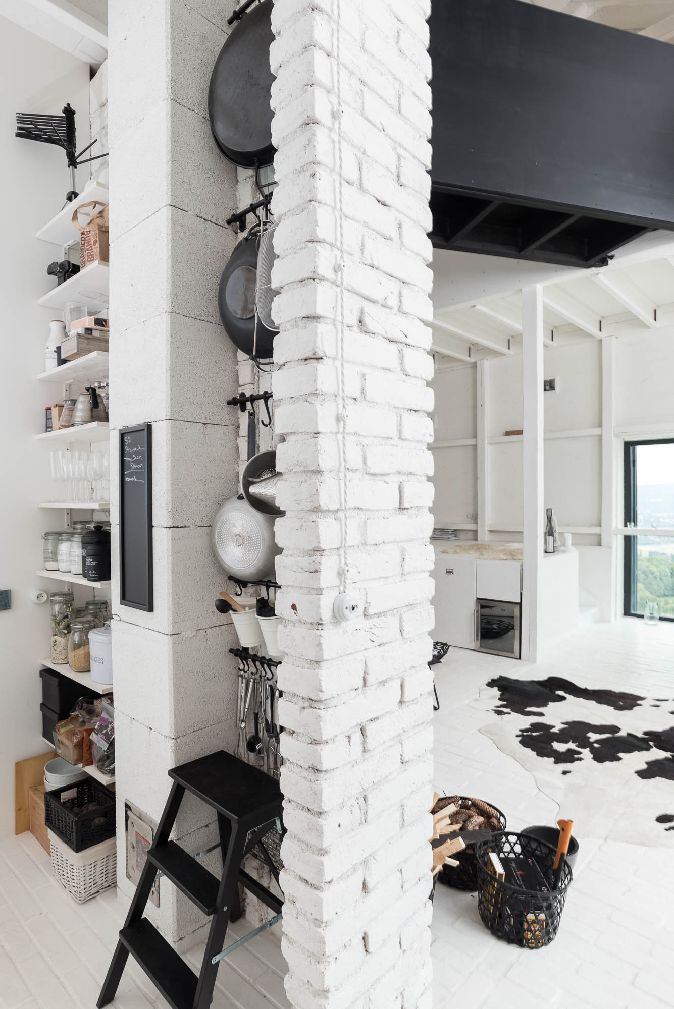 BOX barn kitchen white painted brick pillar — hanging pots and pans on hooks, open shelves with kitchen items, cowhide rug in living area beyond | OOOOX