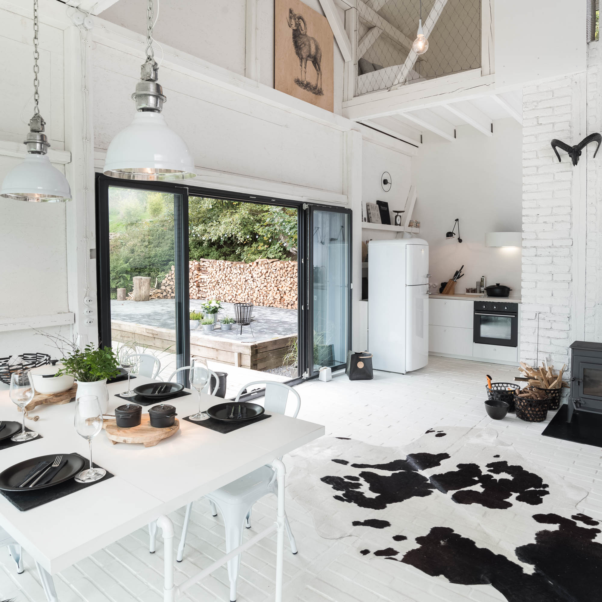 BOX barn interior from dining area — bi-fold glass terrace doors, white retro fridge, upper gallery with ram drawing, wood stove in corner | OOOOX
