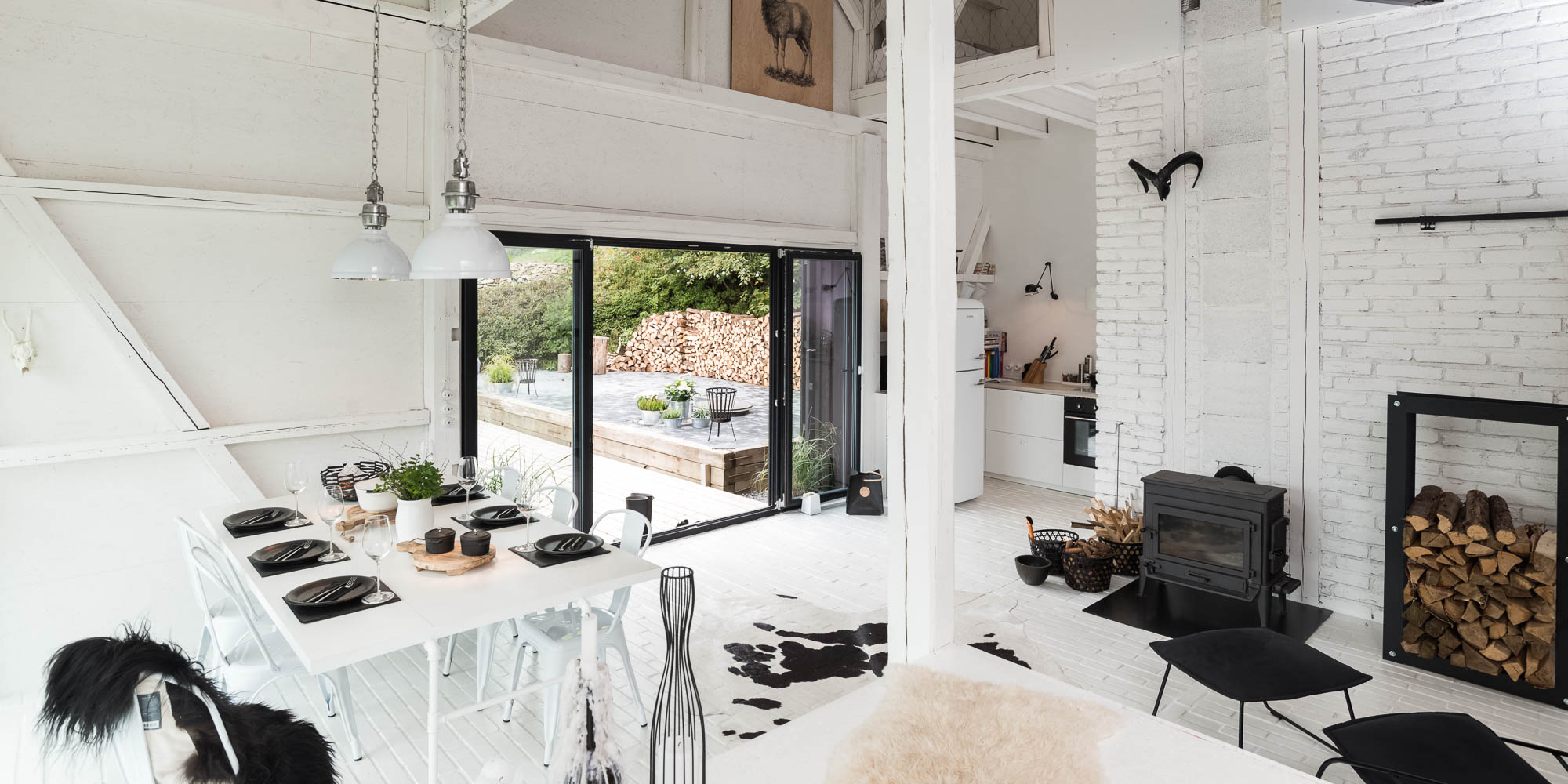 BOX barn interior panorama — dining area, bi-fold terrace doors, kitchen corner, wood stove with firewood, cowhide rug, black lounge chairs | OOOOX