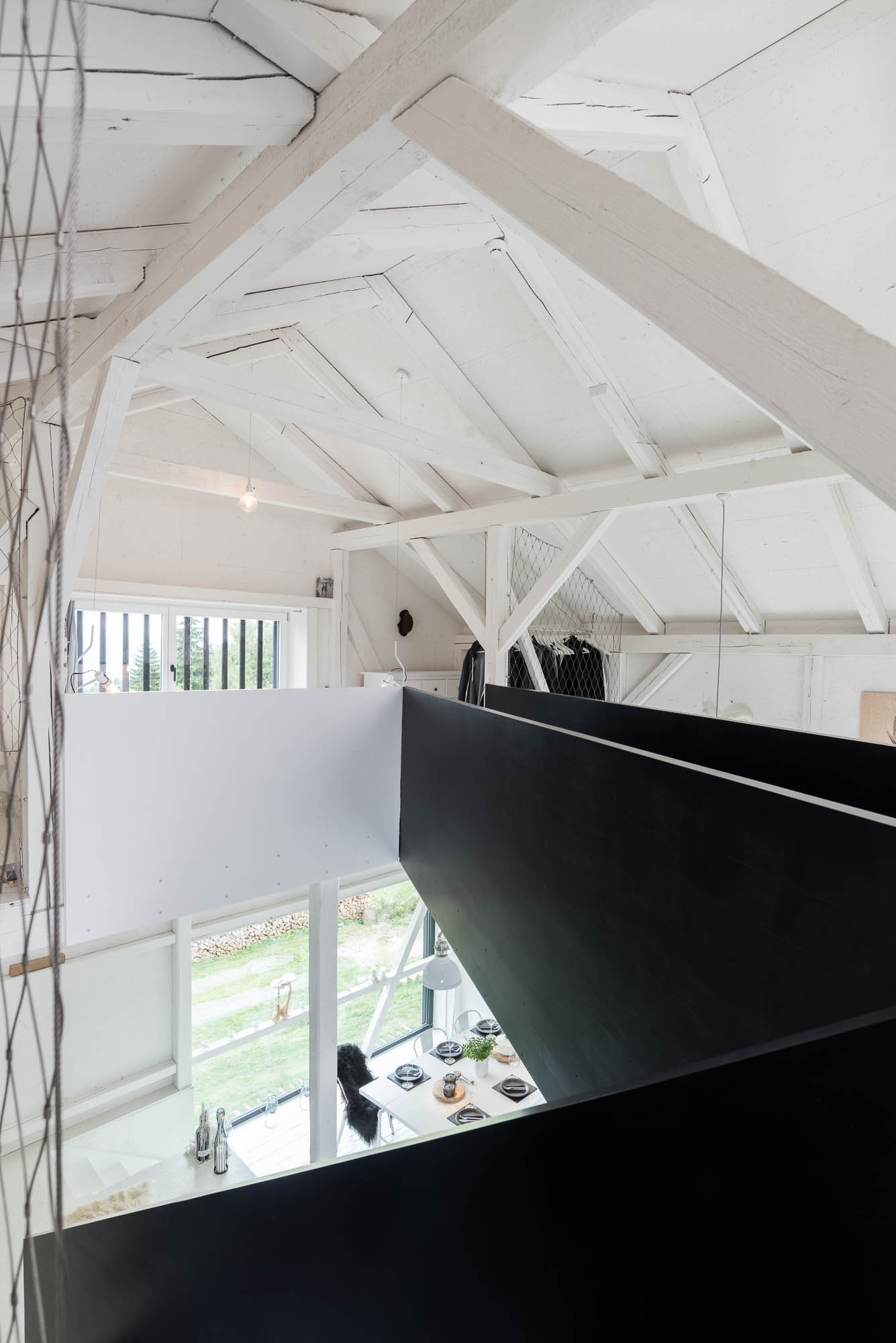 BOX barn view from mezzanine gallery — black steel parapet, white roof trusses, dining table on ground floor below, suspended wire mesh railing | OOOOX