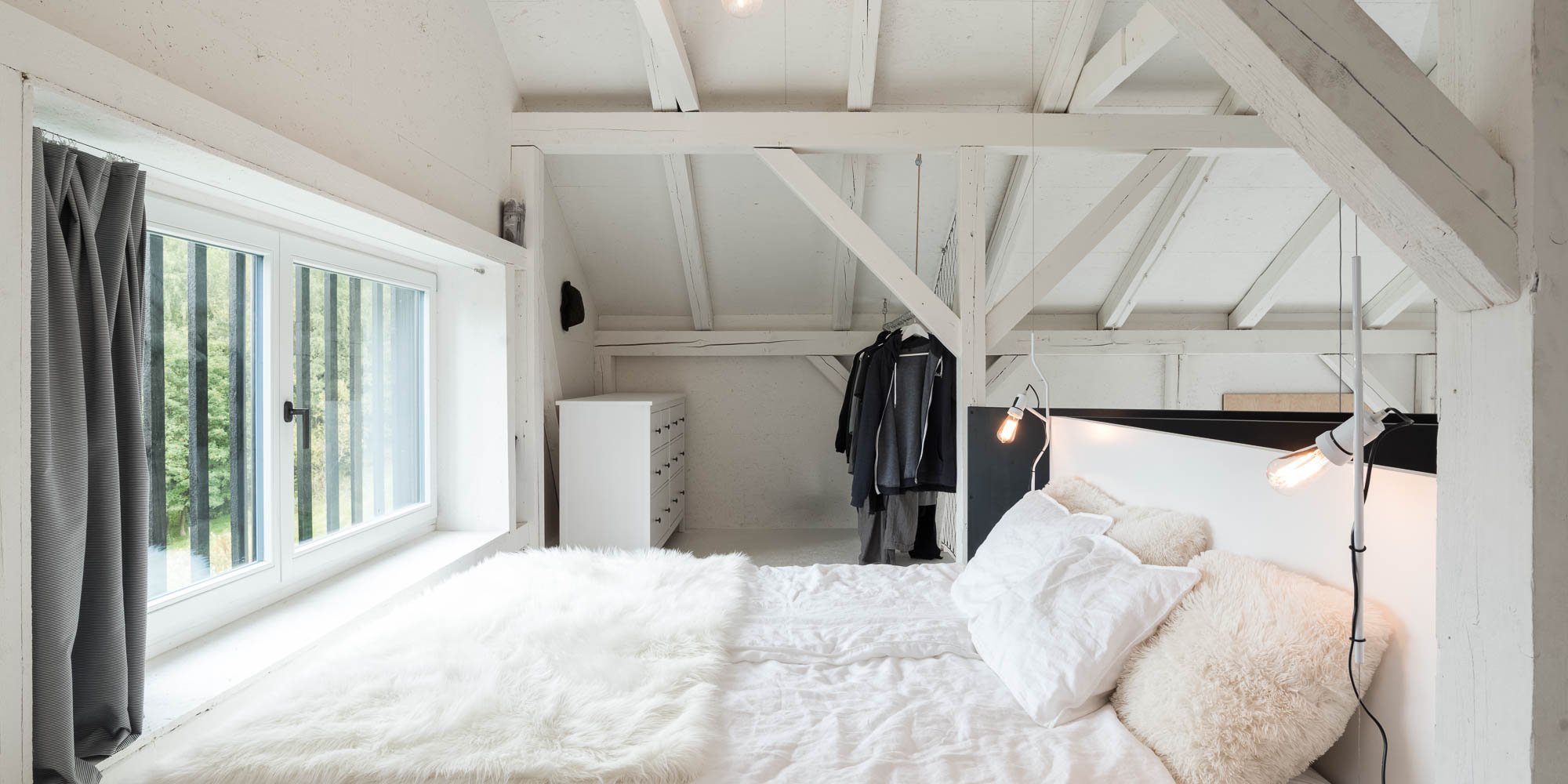 BOX barn attic bedroom — double bed with white linen throw and grey linen pillows, black headboard panel, exposed white timber trusses, window overlooking forest | OOOOX