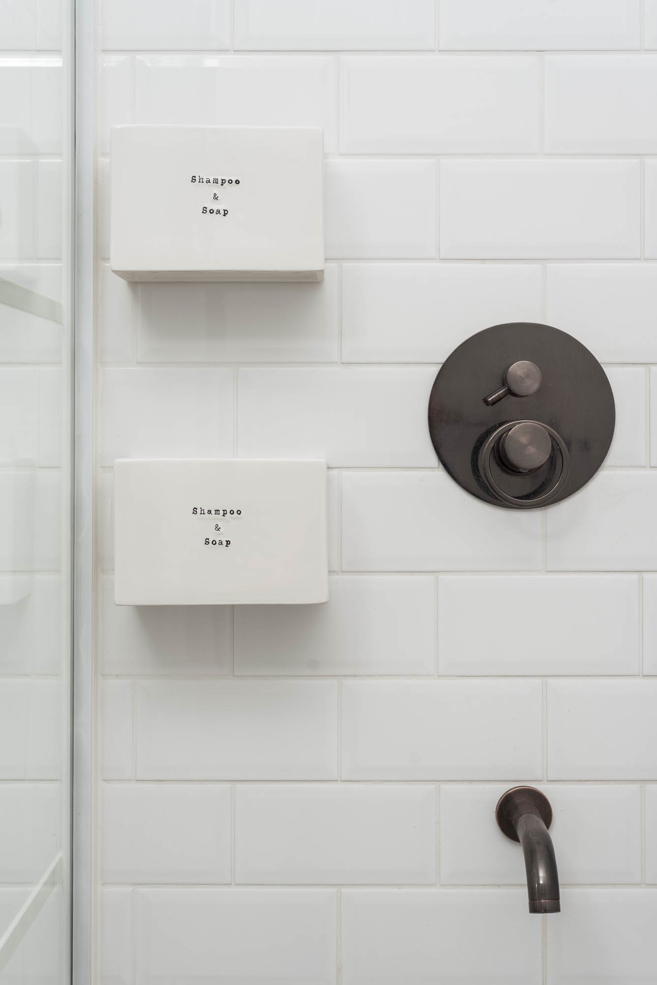 BOX barn shower interior detail — wall-mounted shampoo and soap dispensers, dark bronze thermostatic mixer, bath spout, white subway tiles | OOOOX