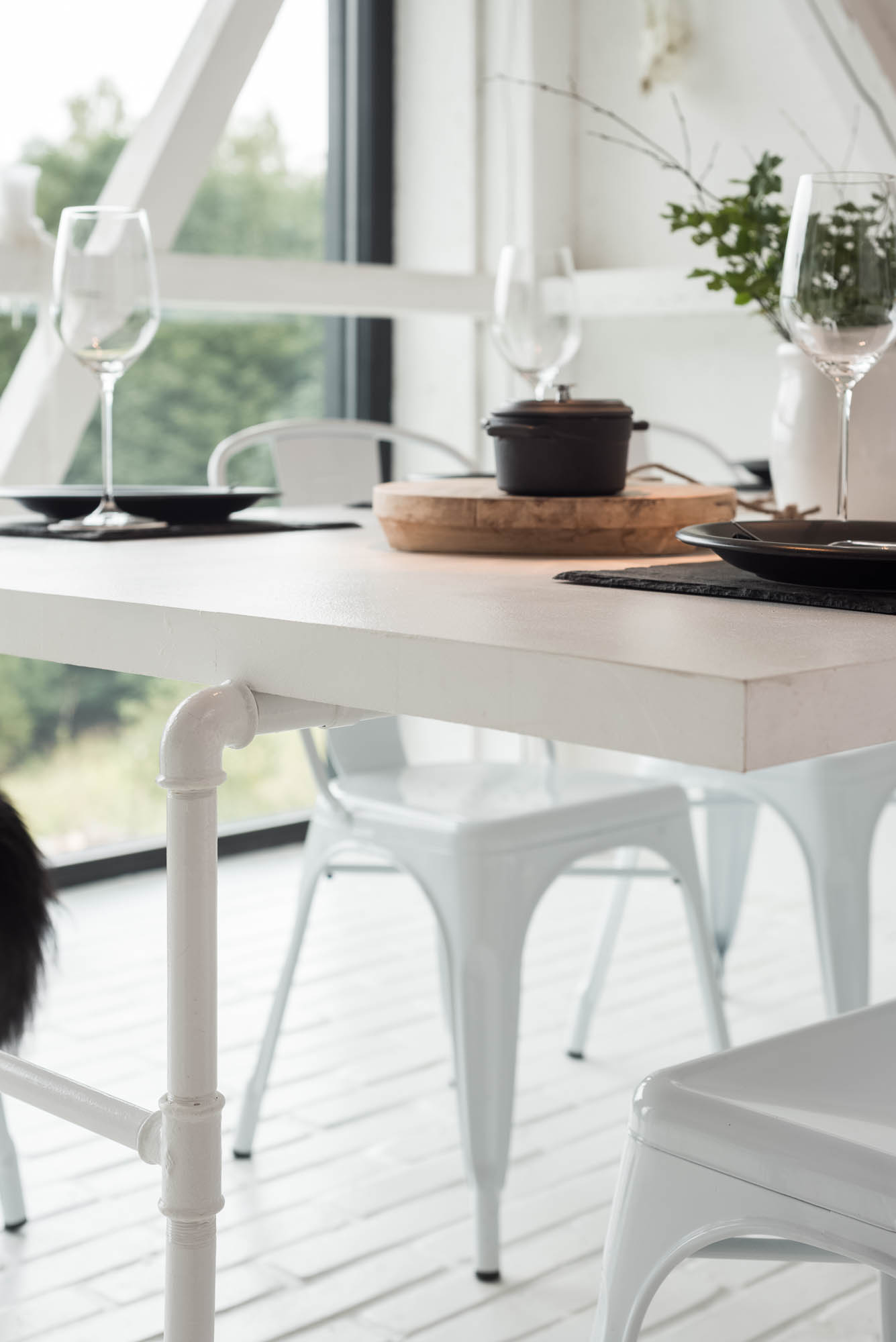 BOX barn dining table detail — white table top on plumbing pipe base leg, black place settings with cutlery, white metal Tolix chairs, arched window to garden | OOOOX