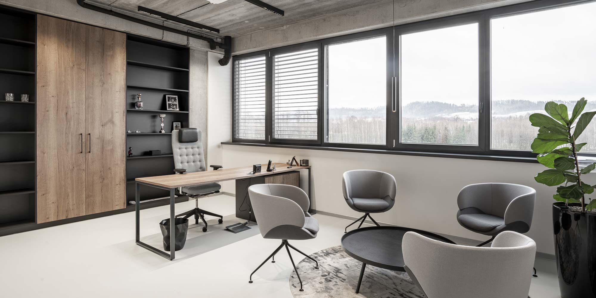Interiér moderní kanceláře s vysokými skříňkami, pracovní stůl s designovým kancelářským křeslem a kruhový stůl s čalouněnými křesly. Betonová stěrka na podlaze a stěnách, přiznané technické potrubí na stropě. OOOOX