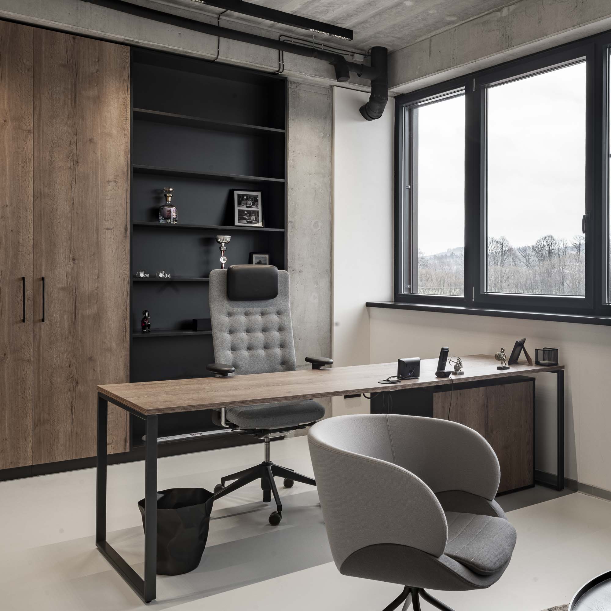 Interiér moderní kanceláře s vysokými skříňkami, pracovní stůl s designovým kancelářským křeslem a kruhový stůl s čalouněnými křesly. Betonová stěrka na podlaze a stěnách, přiznané technické potrubí na stropě. OOOOX