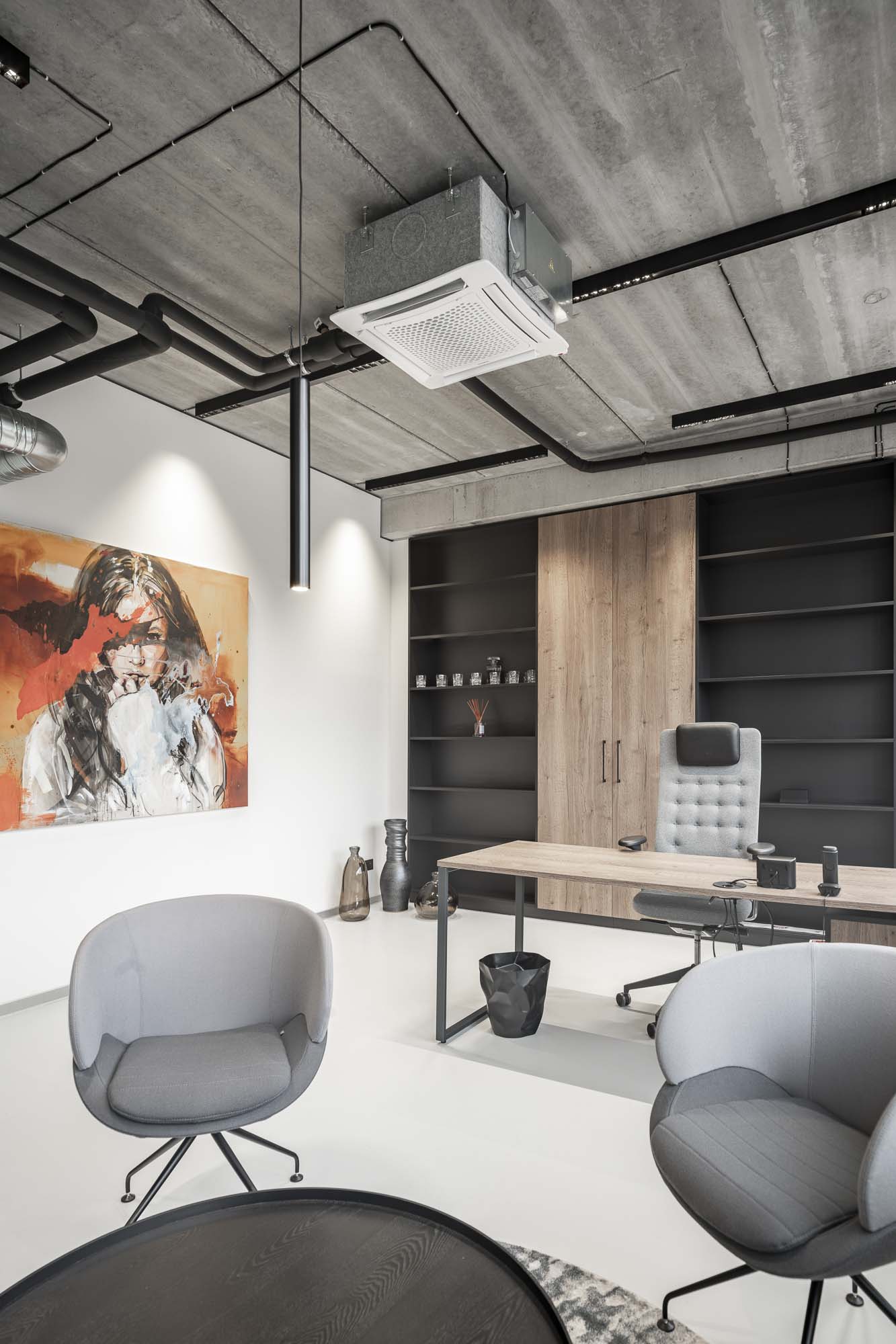 Interiér moderní kanceláře s vysokými skříňkami, pracovní stůl s designovým kancelářským křeslem a kruhový stůl s čalouněnými křesly. Betonová stěrka na podlaze a stěnách, přiznané technické potrubí na stropě. OOOOX