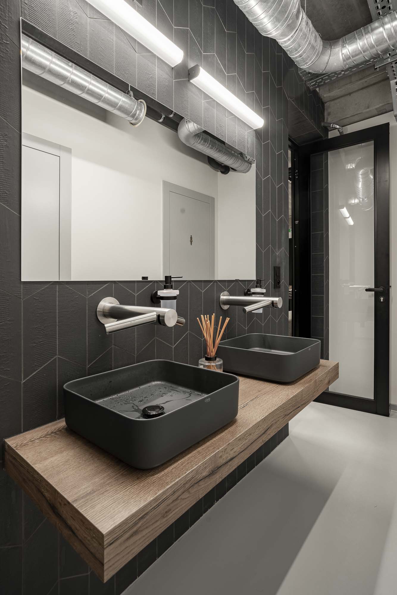 Moderní umývárna a toalety v industriálním stylu s betonovou stěrkou, černými detaily, minimalistickým osvětlením a velkoformátovým obkladem. Design by OOOOX