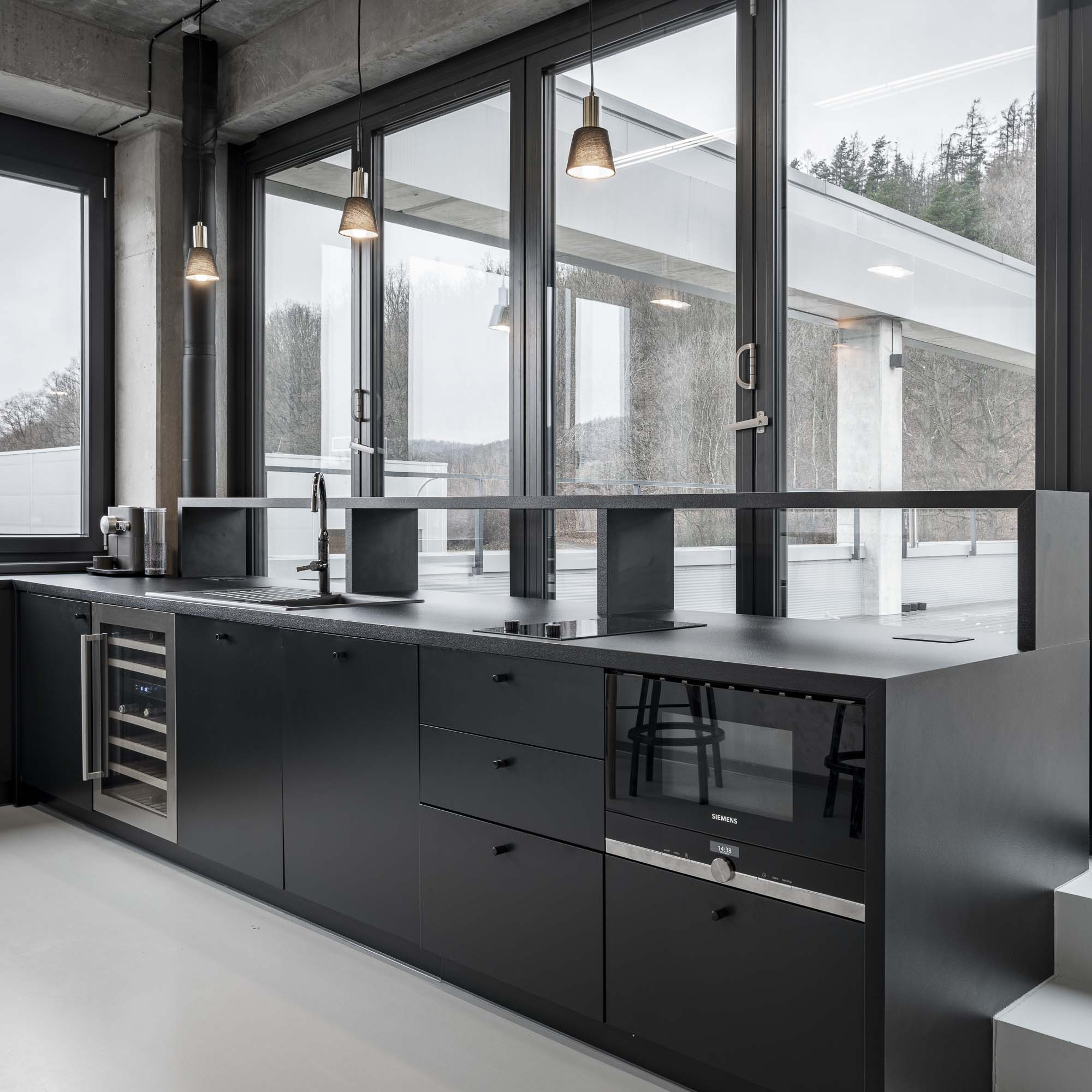 Minimalistická kancelářská kuchyňka v černém odstínu, betonovými povrchy a vestavěným nábytkem na míru v industriálním interiéru kanceláří. Design by OOOOX