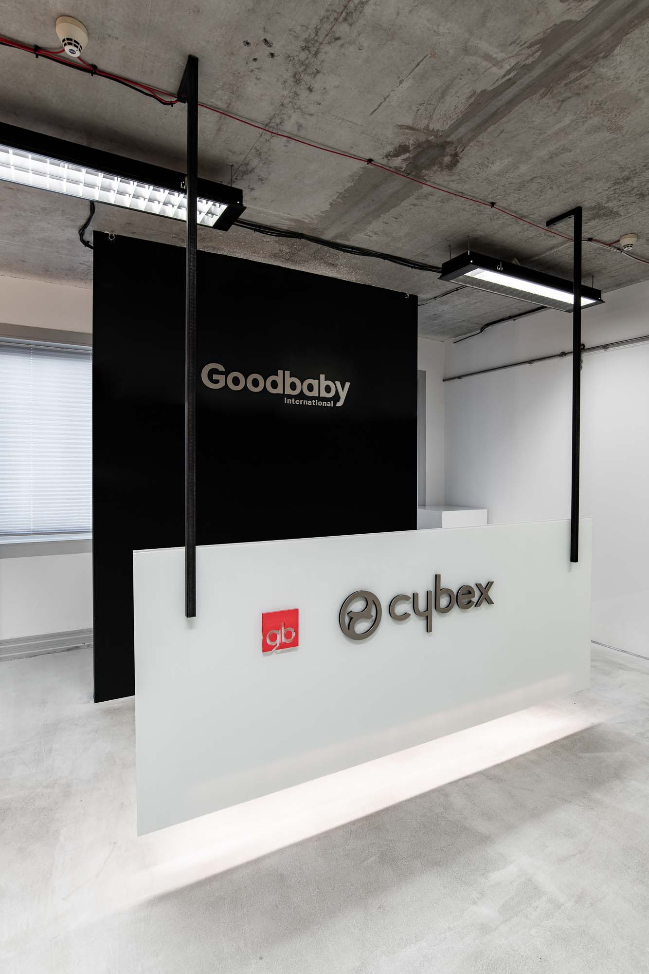 Recepce kanceláří Cybex Goodbaby Praha; plovoucí bílý recepční pult s logem Cybex a červeným logem gb na čelním panelu, velký černý panel s nápisem Goodbaby International za pultem, dva černé ocelové sloupy