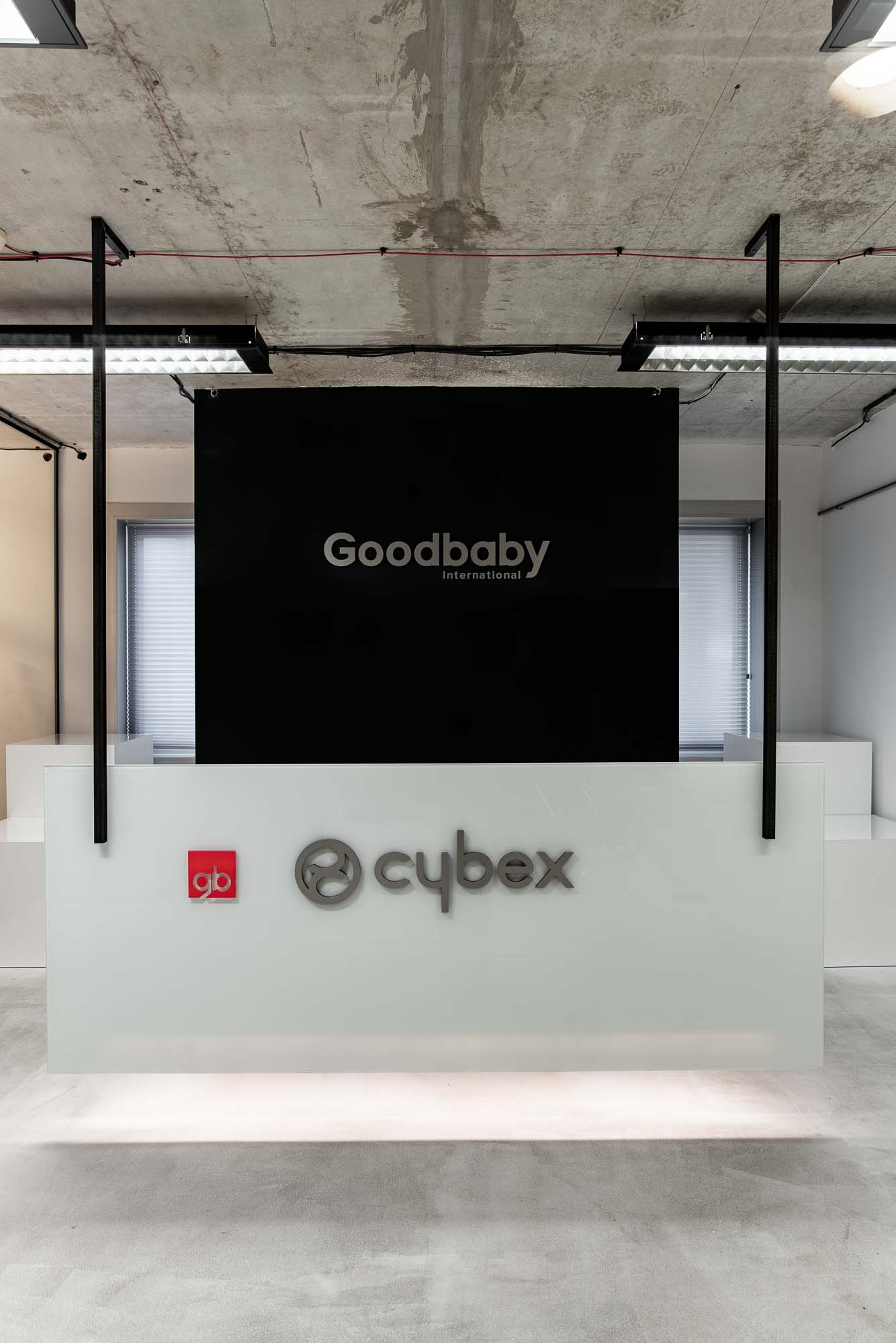 Čelní pohled na recepční pult kanceláří Cybex Goodbaby Praha; bílý plovoucí pult s logy Cybex a gb, vyvýšený černý panel s nápisem Goodbaby International ve stříbrných písmenech, okna s žaluziemi po stranách, leštěná betonová podlaha