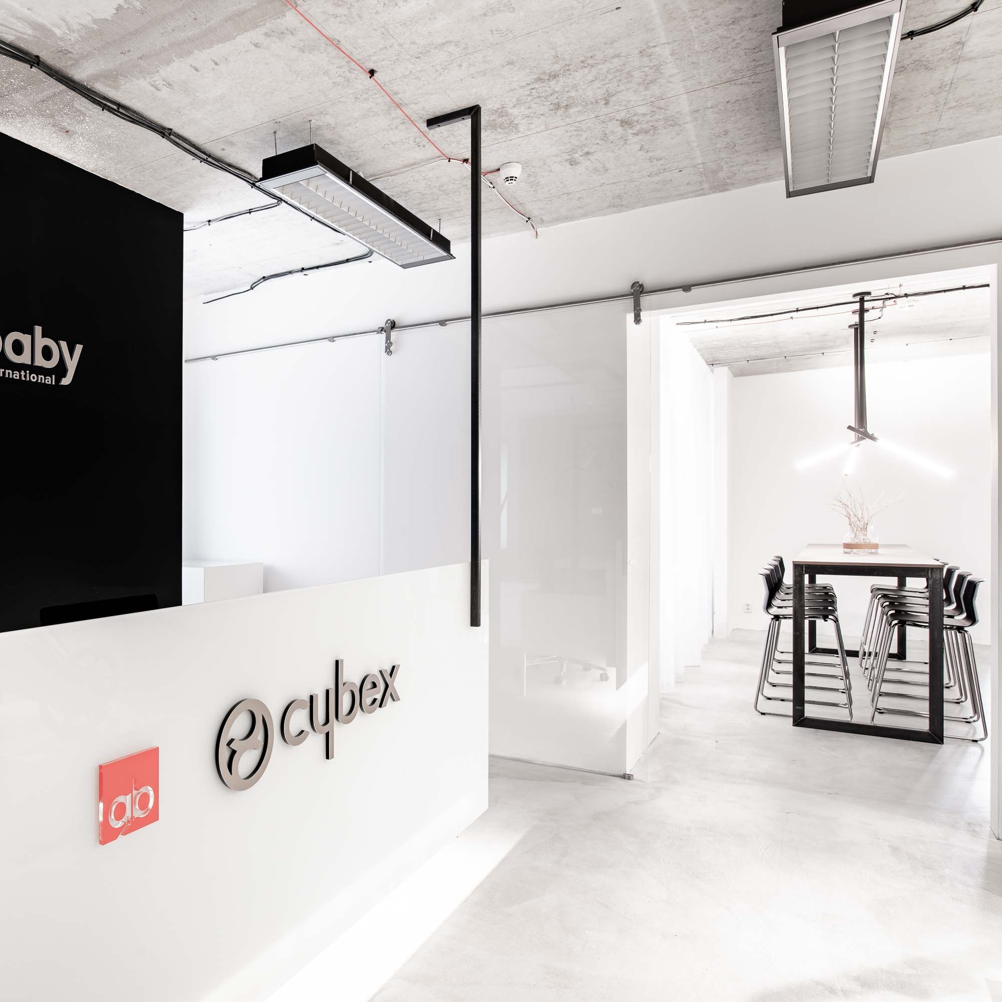 Pohled z recepce Cybex Goodbaby směrem k jídelní a společenské zóně; bílý pult s logem Cybex, chodba ústící k černému rámovému stolu s dubovou deskou, černé sáňkové židle, dekorativní větvičky ve váze a závěsné světlo – kanceláře Praha