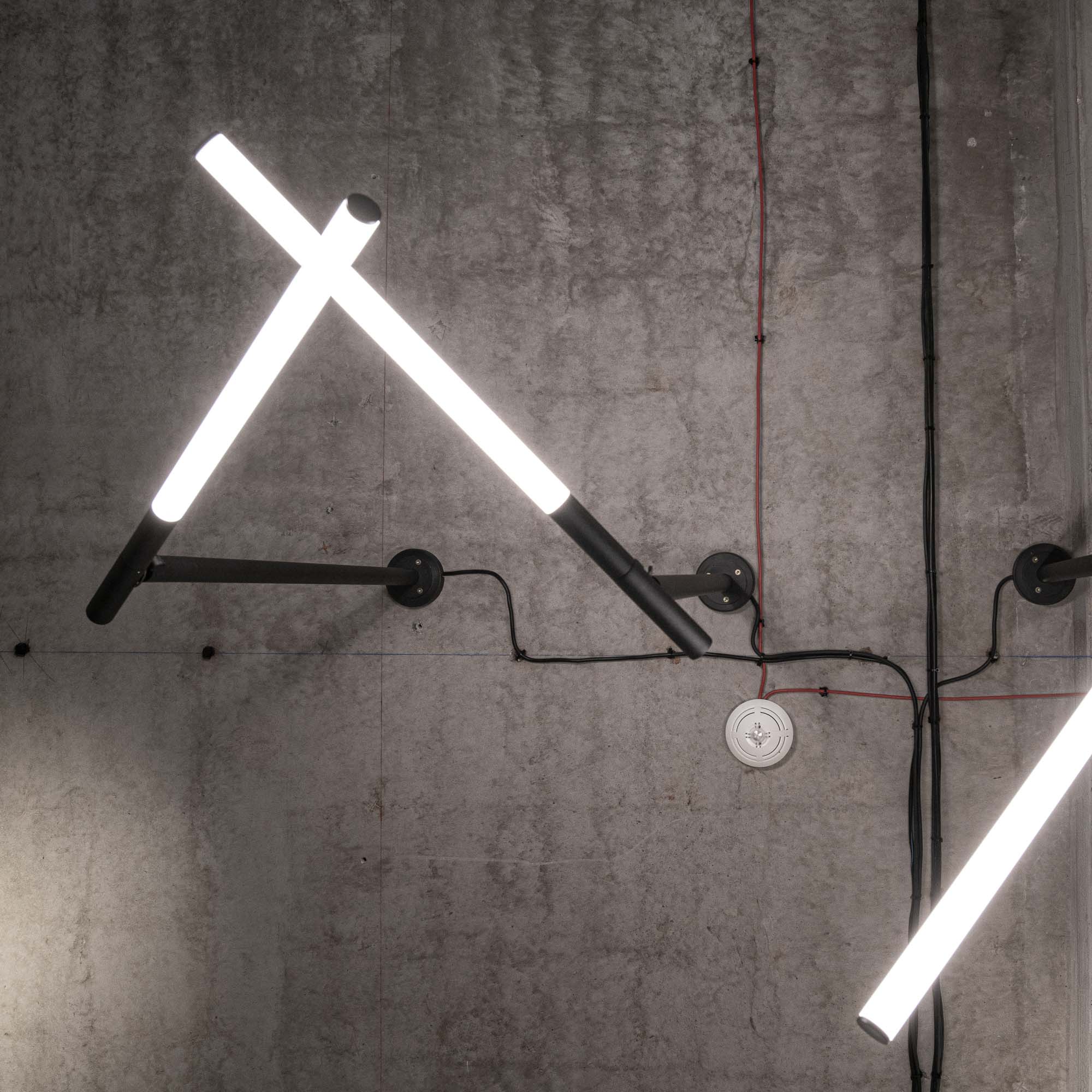 Detail nástěnných geometrických světelných svítidel na pohledové betonové stěně; dvě nastavitelné černé kovové ramena s křížícími se LED trubicemi tvořícími vzor X, upevněné na betonovou stěnu černými trubkovými úchyty