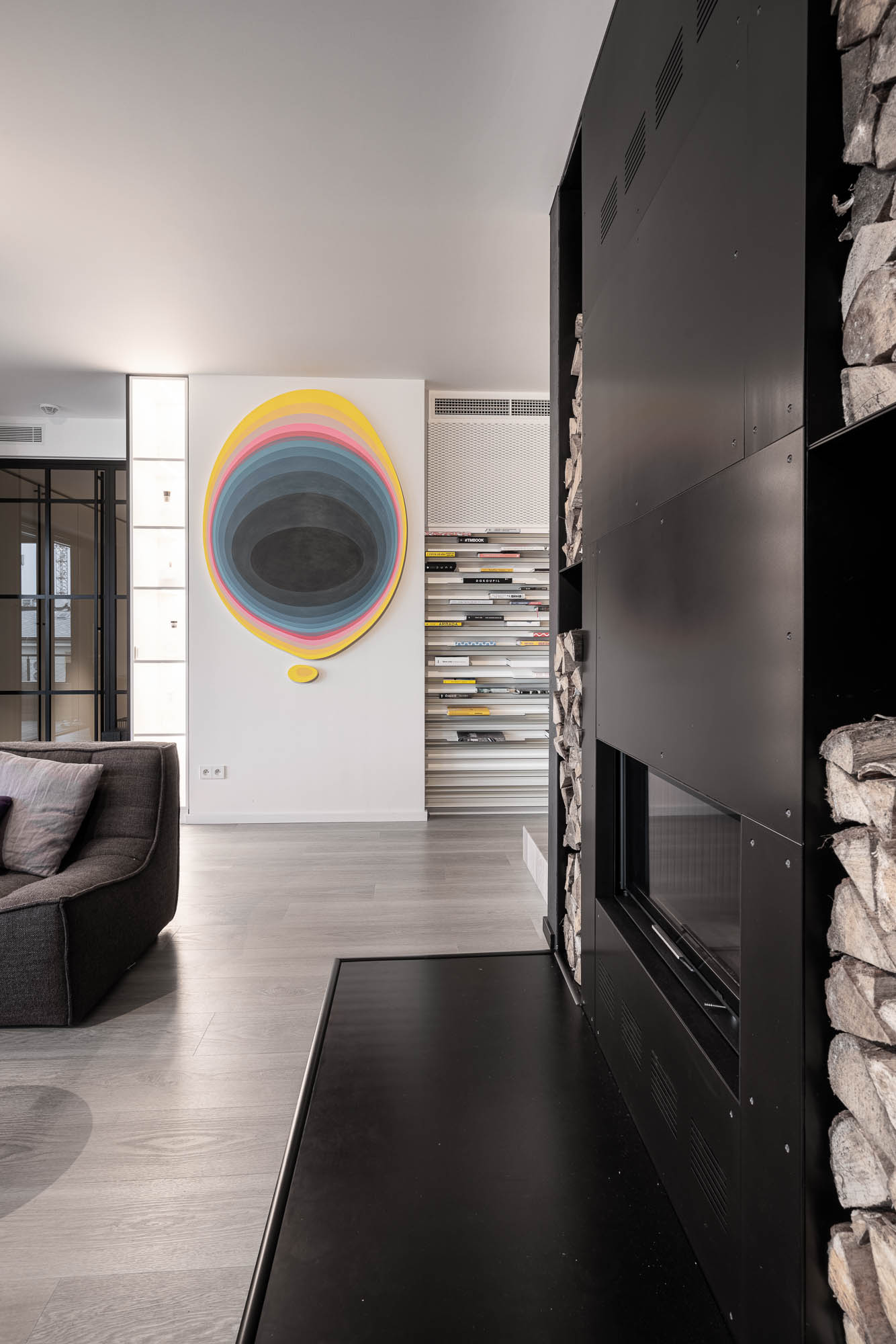 Obytný prostor mezonetového bytu s výrazným žlutým schodištěm a černým krbem, Black steel fireplace log niche magazine display shelf colourful oval painting minimalist interior OOOOX Praha