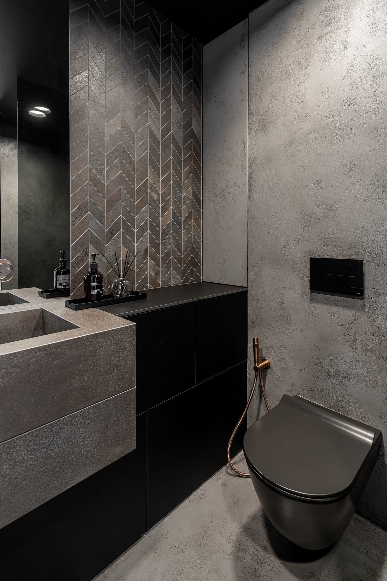 Detail koupelny vily Hřebenky – šikmá tmavá mozaika, betonový umyvadlový vanity, černá závěsná mísa, mosazná hygienická sprcha, tmavá atmosféra