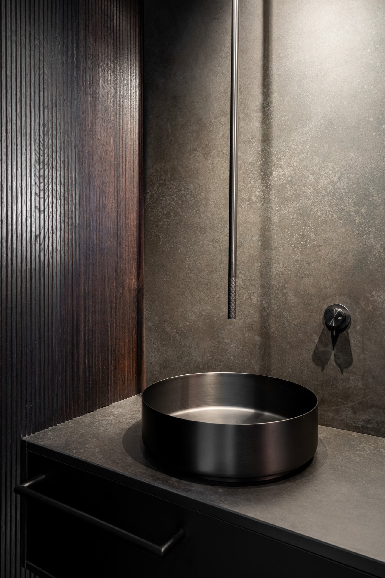 Detail umyvadla WC vily Hřebenky – kulaté černé/gunmetalové umyvadlo na tmavém pultu, úzké zrcadlo s LED páskem nad ním, rýhovaný tmavý dřevěný panel, šedá minerální stěna