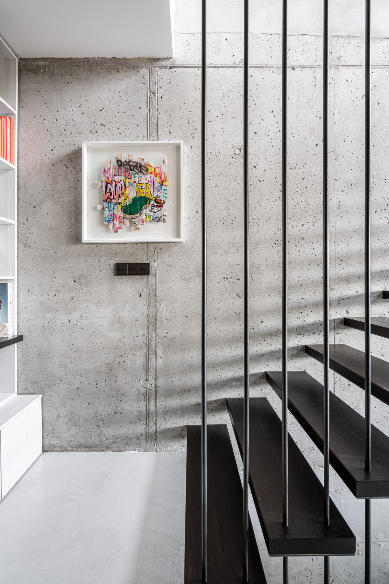 Detail schodiště vily Hřebenky s uměním – pohledový beton, ocelová tyčová příčka, černé ocelové stupně, rámovaný barevný pop-artový obraz s graffiti motivem na betonové stěně, v pozadí knihovna