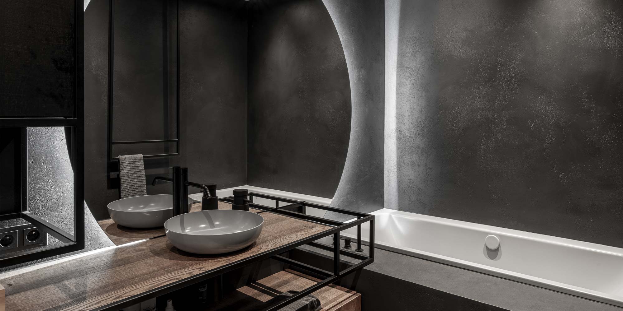 Minimalist bathroom with concrete microcement on floors and walls, circular backlit mirror and black steel accessories. Design by OOOOX