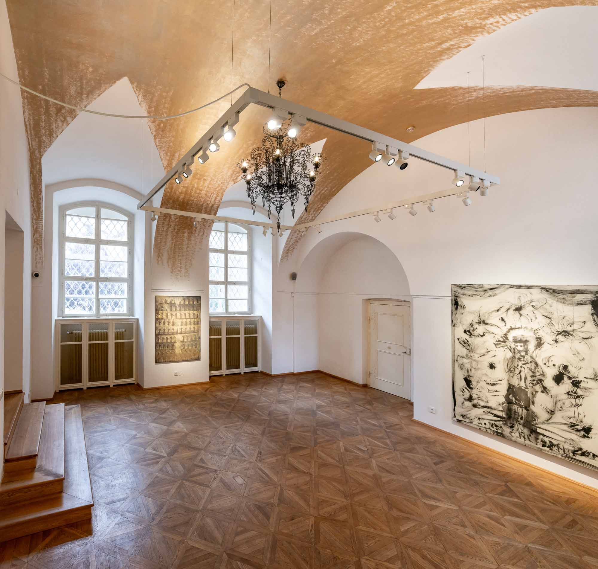 Hlavní výstavní sál galerie ARCIMBOLDO v Praze. Velké drátěné svítidlo s historickým motivem v bílé klenbě a umělecká díla v nikách.